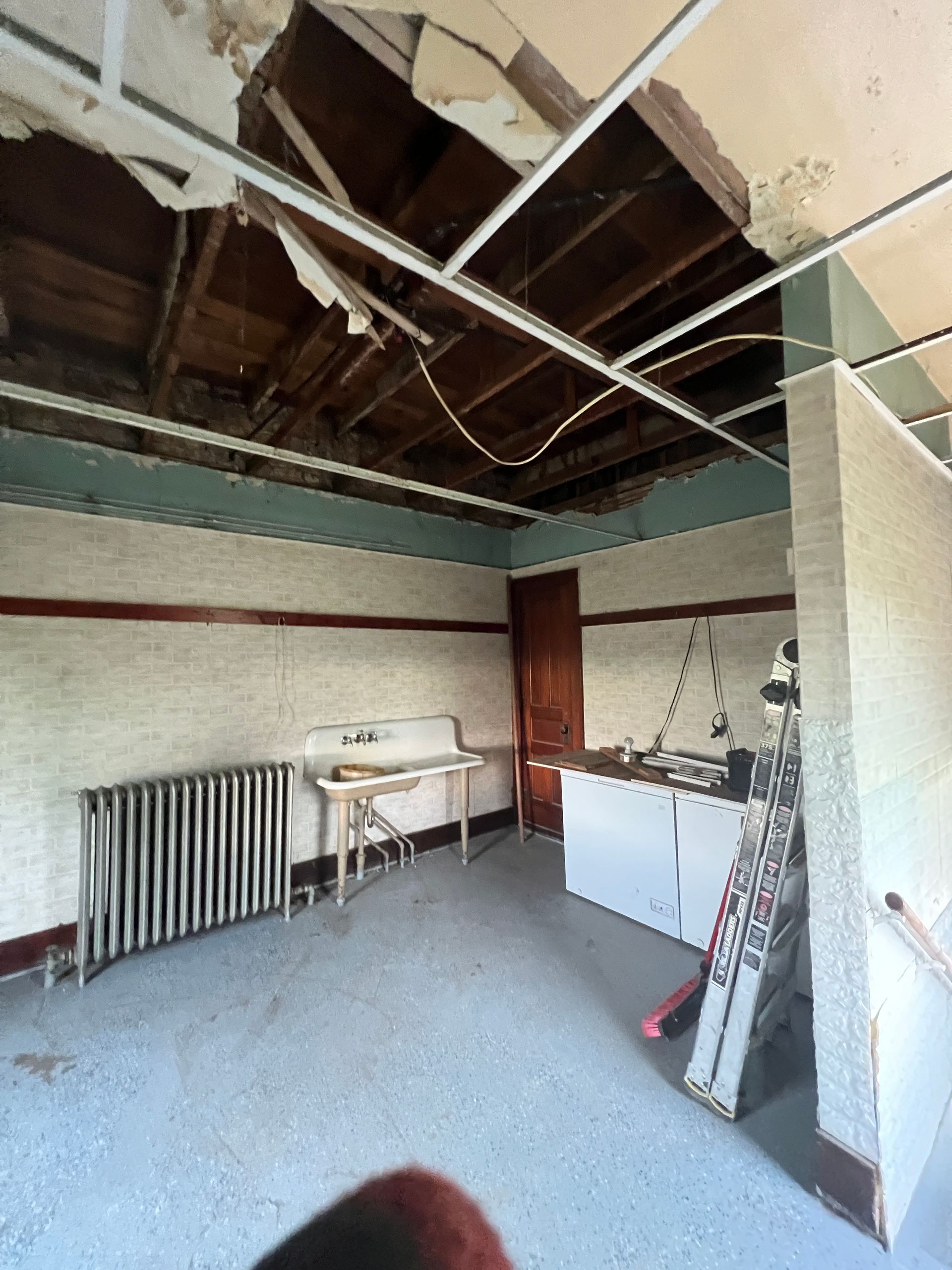 A room under renovation with an exposed ceiling, a radiator, a vintage sink, and a step ladder on a concrete floor.