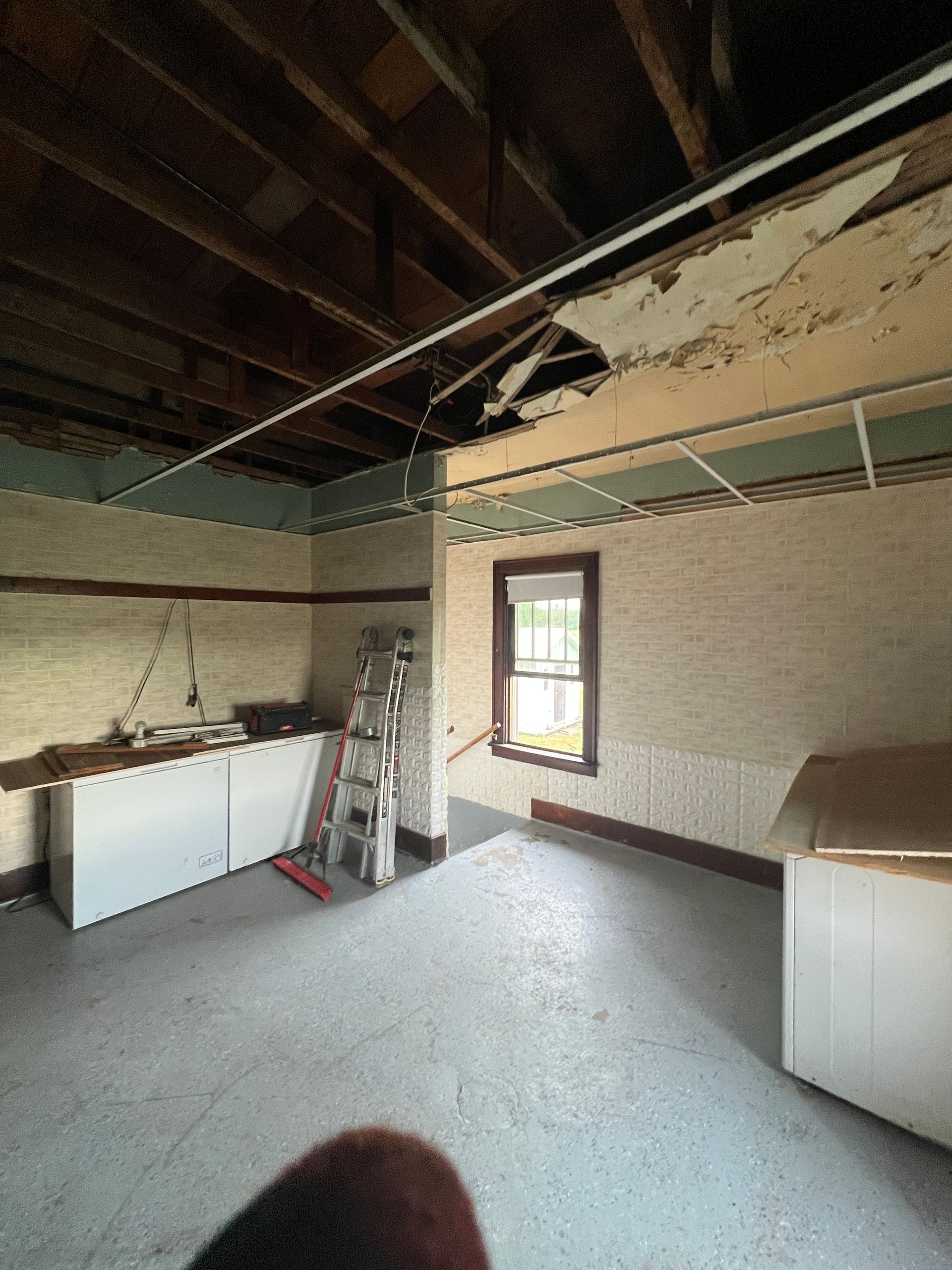 An unfinished room under construction, featuring exposed ceiling joists, damaged drywall, a window, and a ladder.