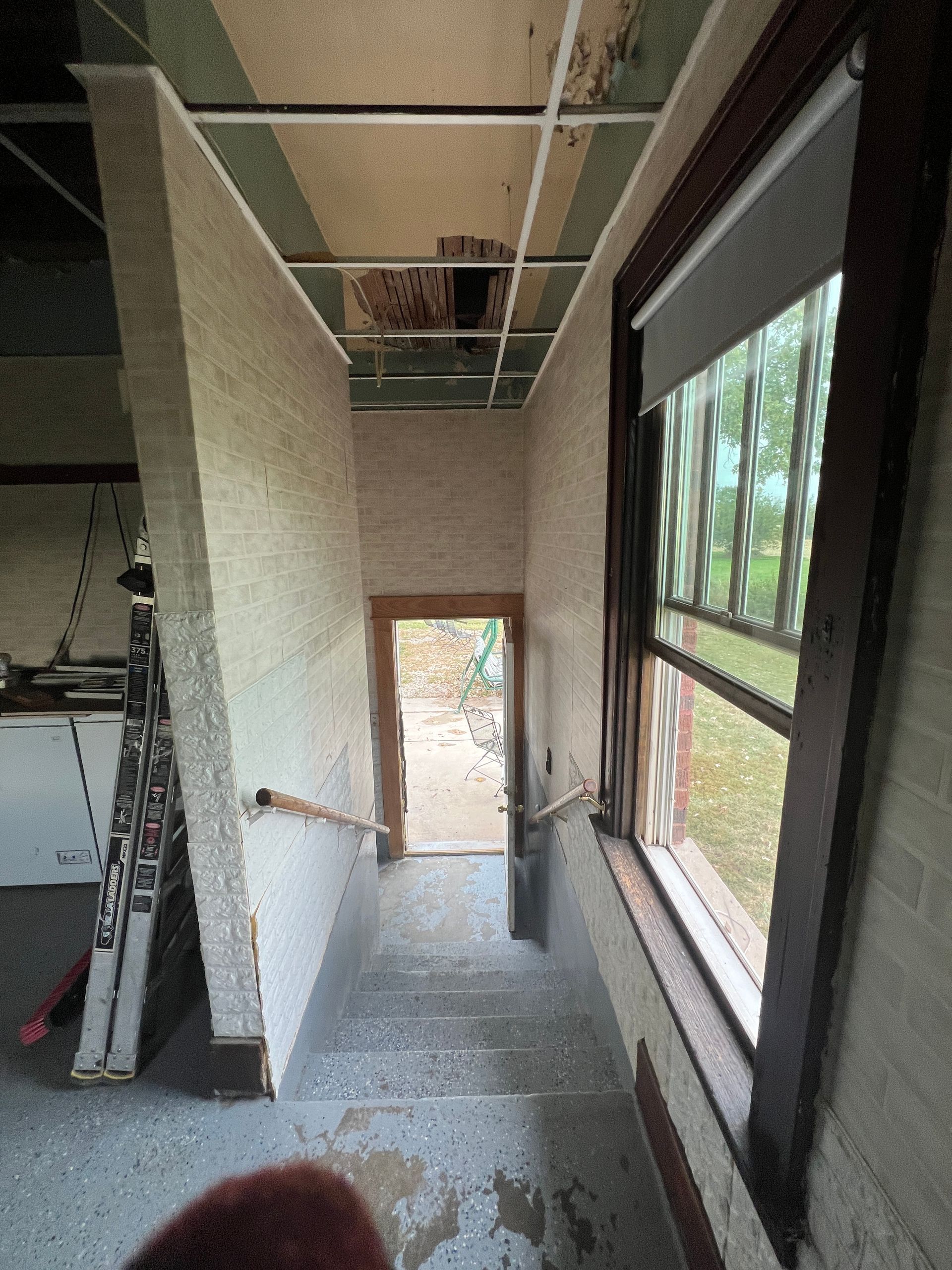 A descending staircase inside a building under renovation, showing a window, a ceiling gap, and an open doorway at the end.