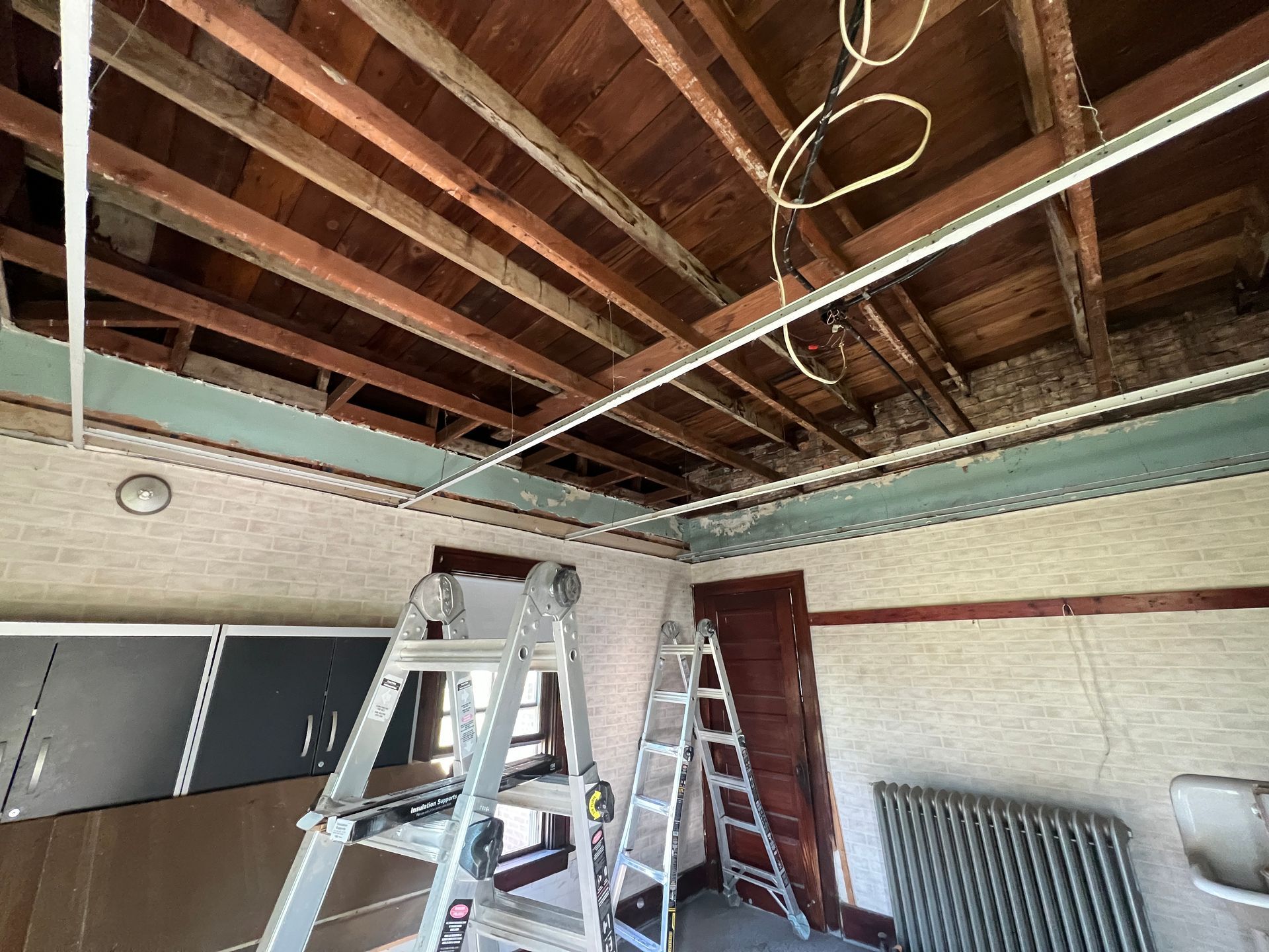 An interior renovation scene showing exposed wooden ceiling beams, two step ladders, and a radiator in a room.