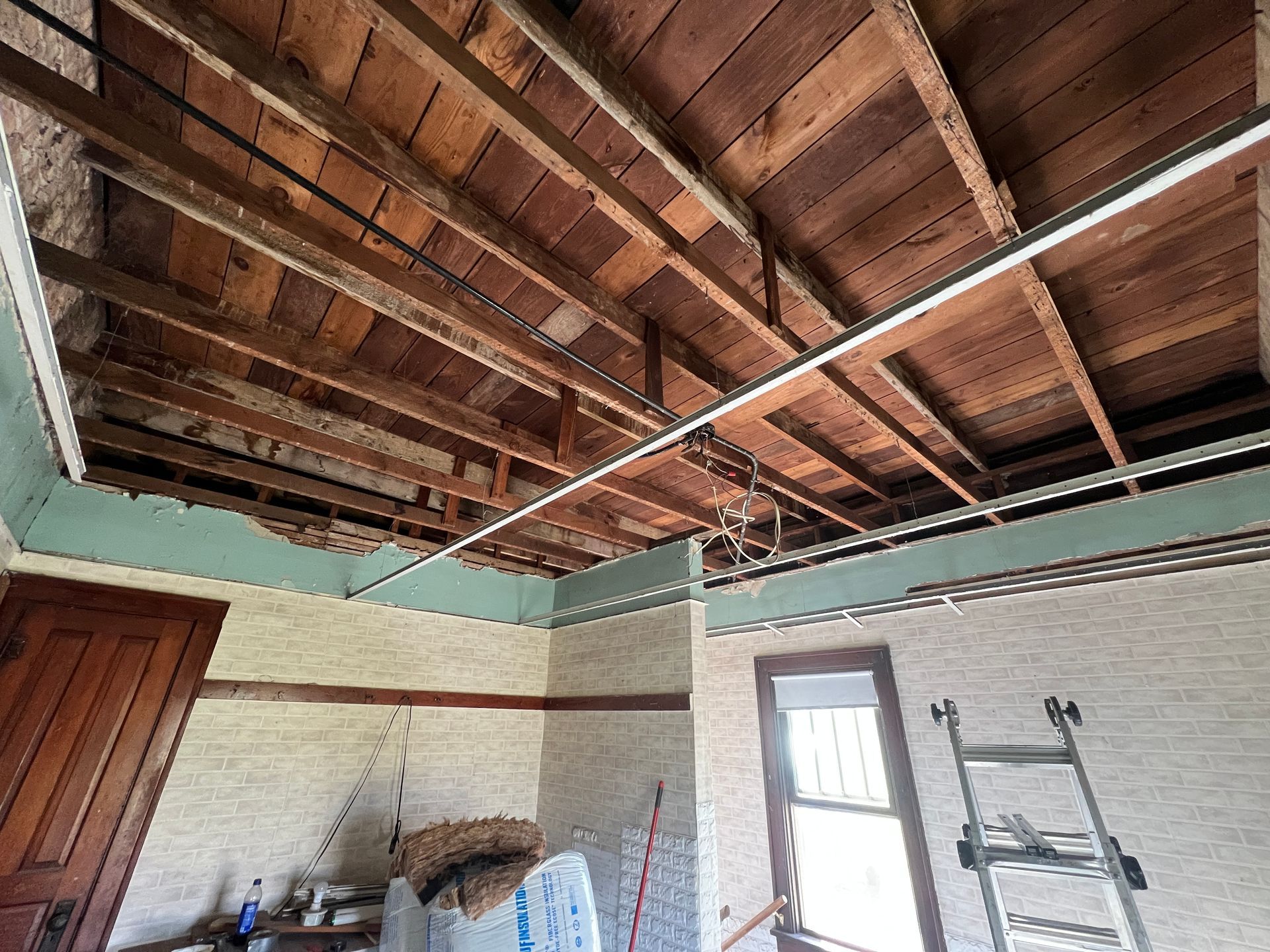 An unfinished room with exposed wooden ceiling joists, teal painted wall trim, a wooden door, and a ladder by a window.
