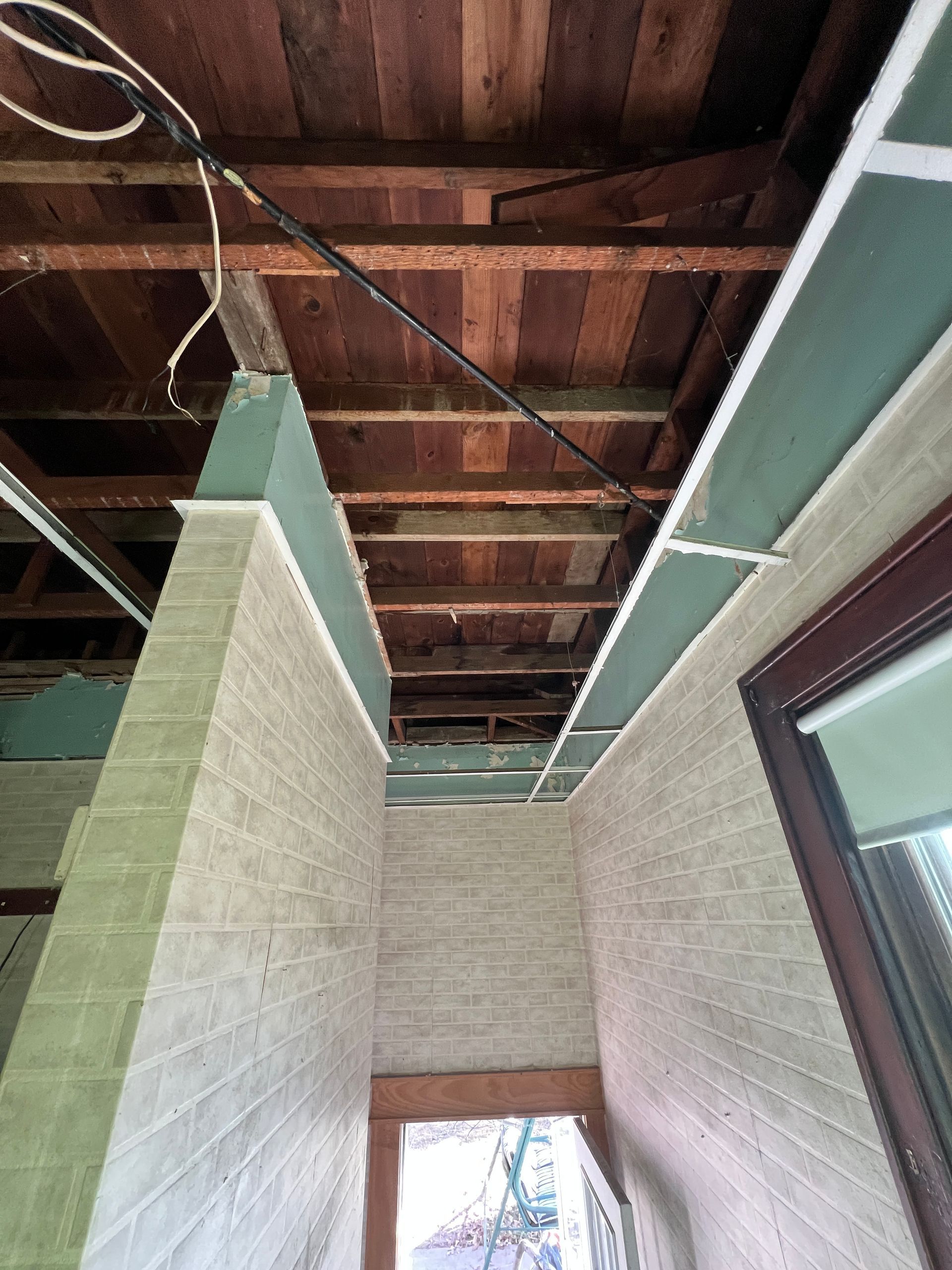 A view looking up at exposed wooden ceiling joists and unfinished drywall above a hallway with textured brick walls.