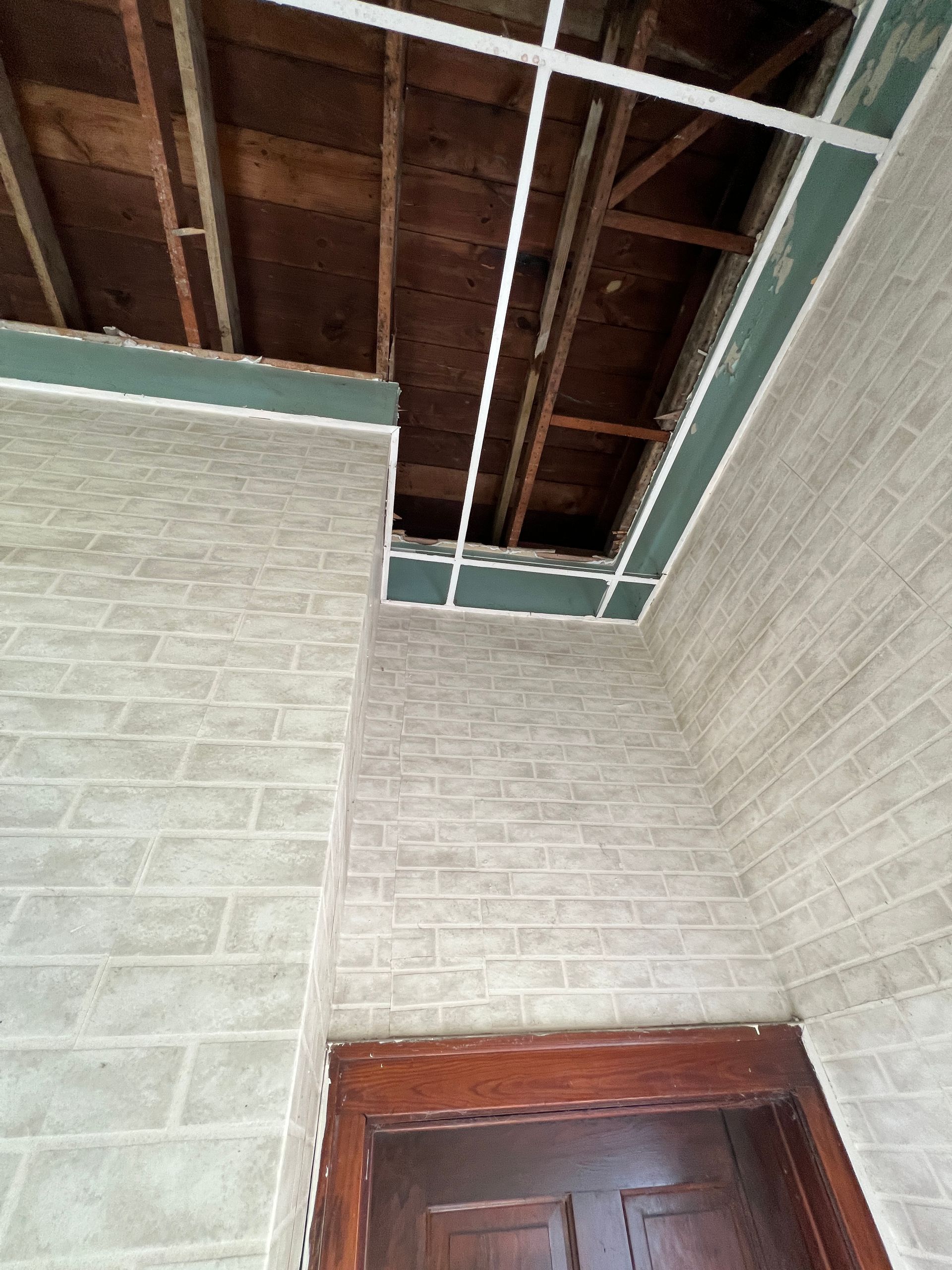 A view looking up at a damaged interior ceiling showing exposed wooden rafters, surrounded by light brick-patterned walls.