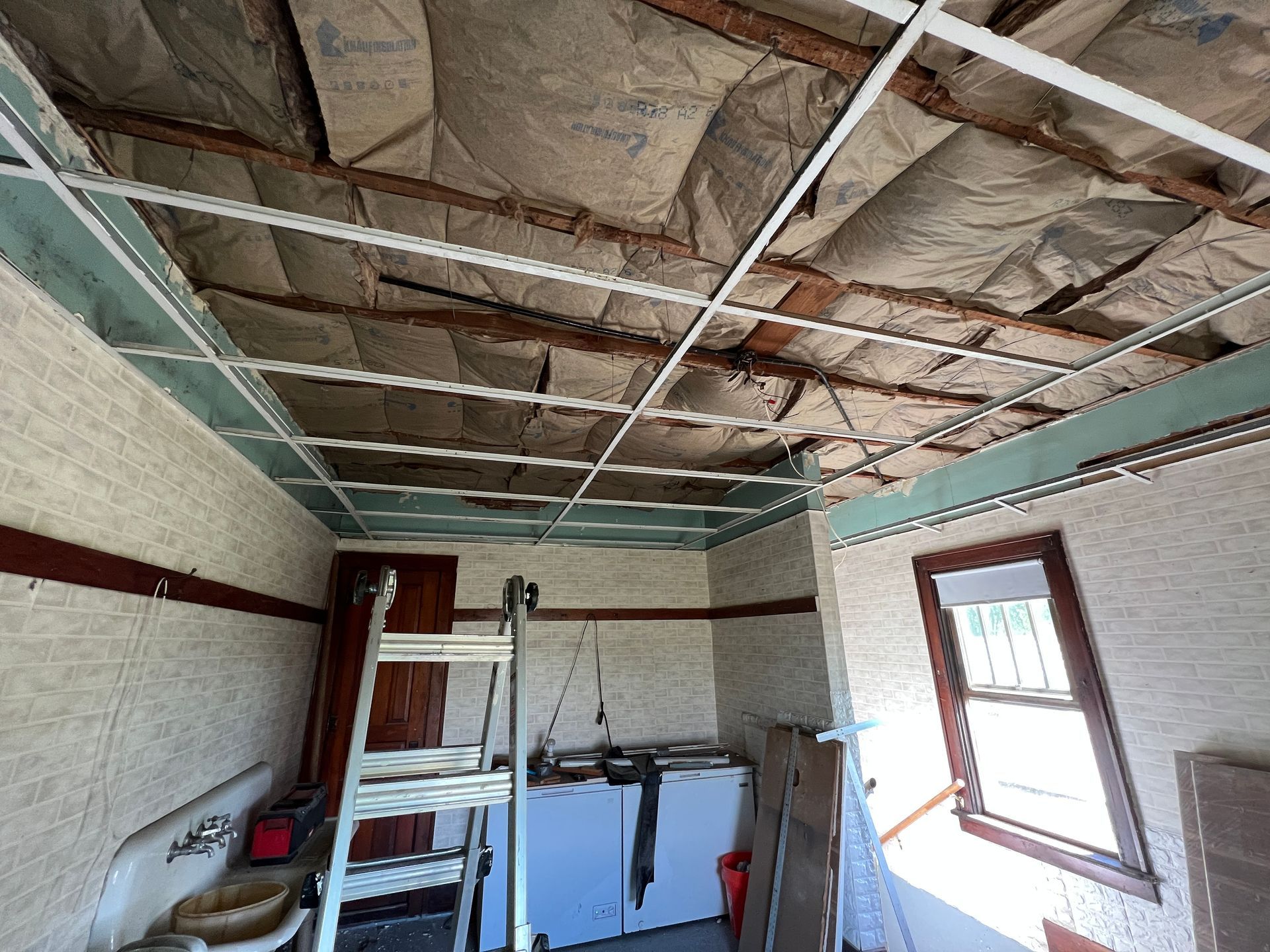 A room under renovation with a suspended metal ceiling grid, exposed insulation, a stepladder, and white appliances.