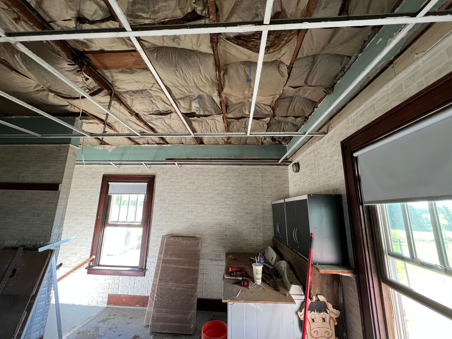 An interior view of a room under renovation with exposed insulation in the ceiling grid and two windows.