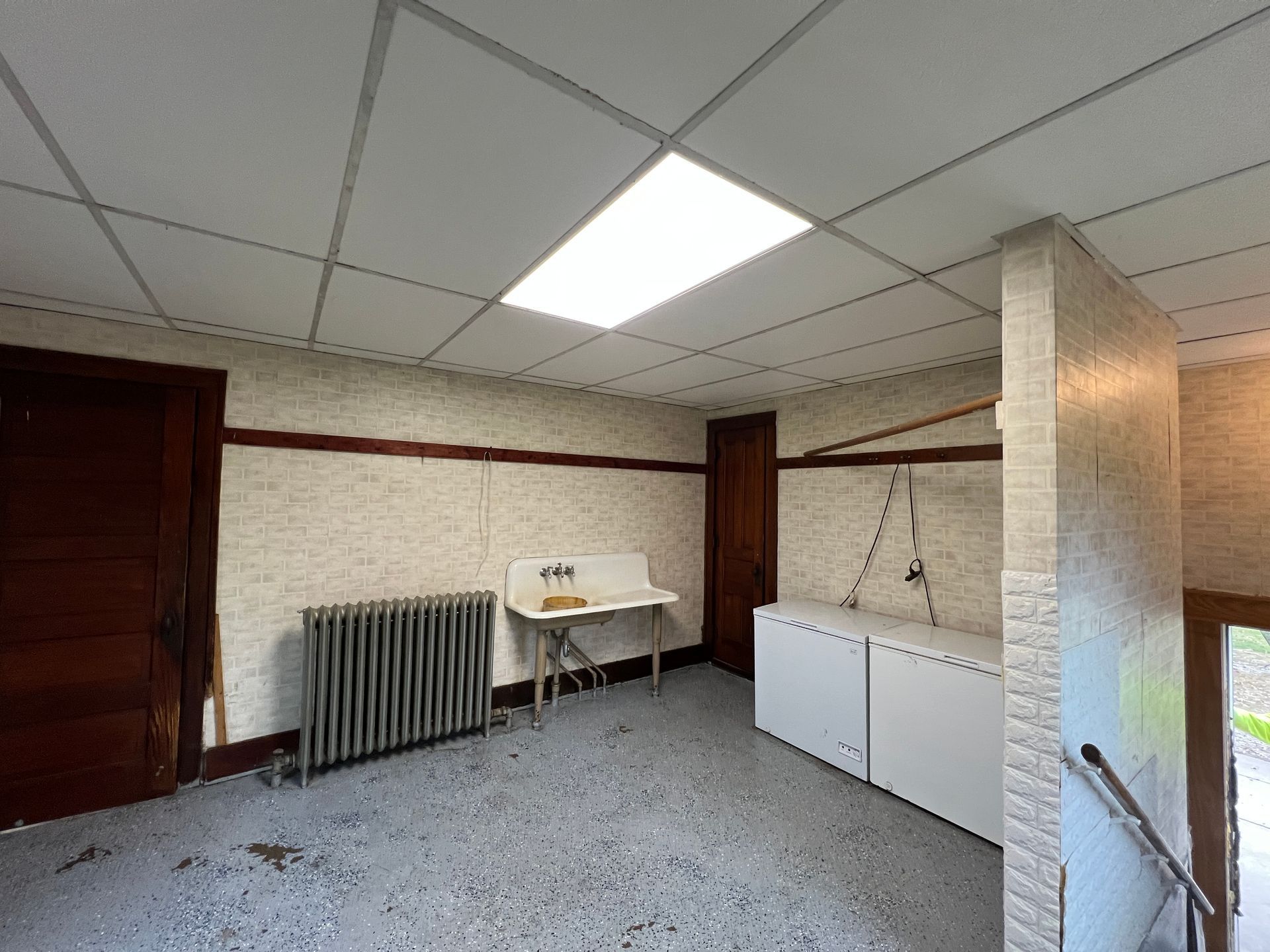 A room with a drop ceiling, beige wallpaper, a radiator, a white utility sink, and two white chest freezers.