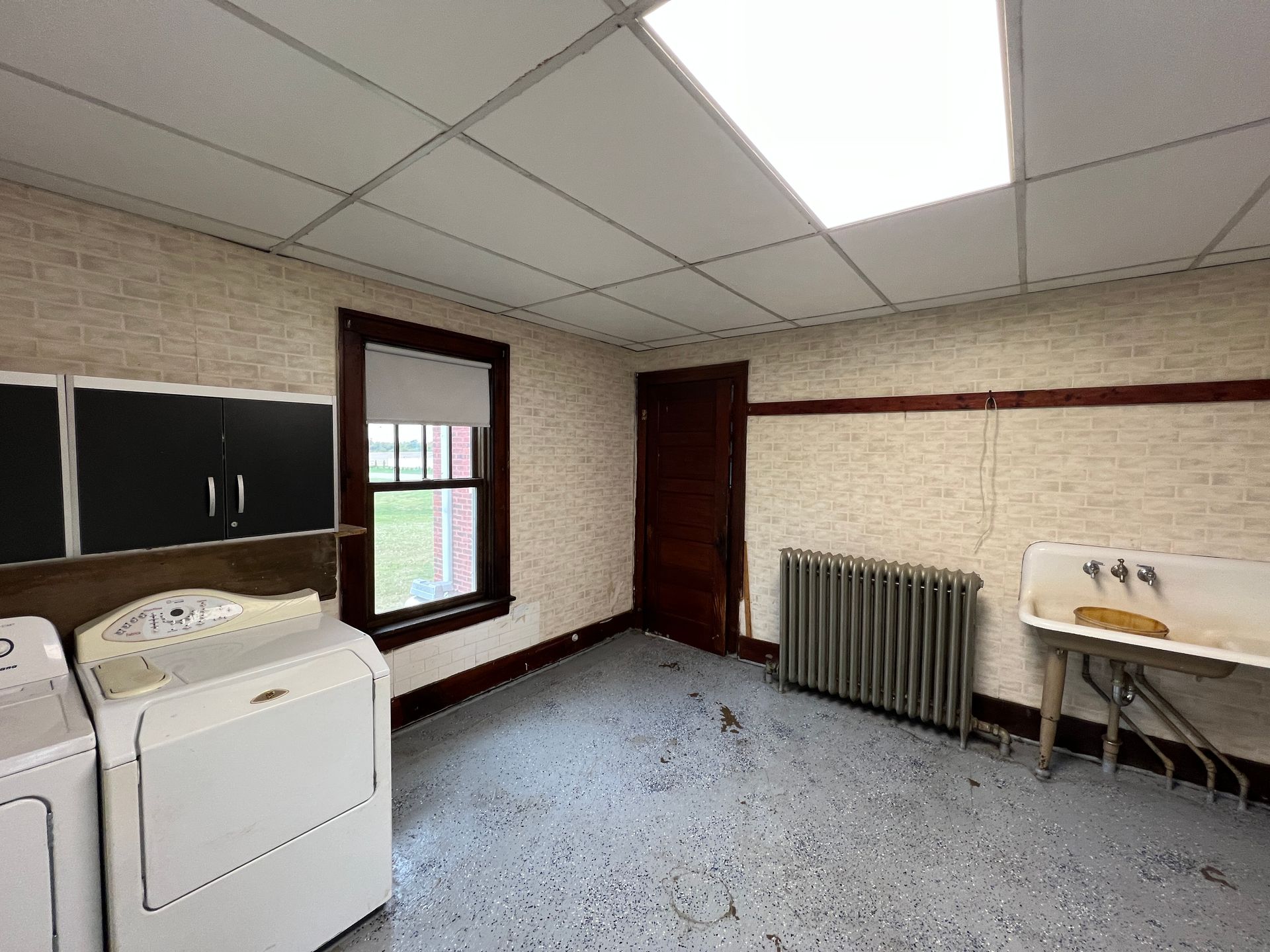 A laundry room with white appliances, dark cabinets, a vintage wall-mounted sink, a radiator, and a window.