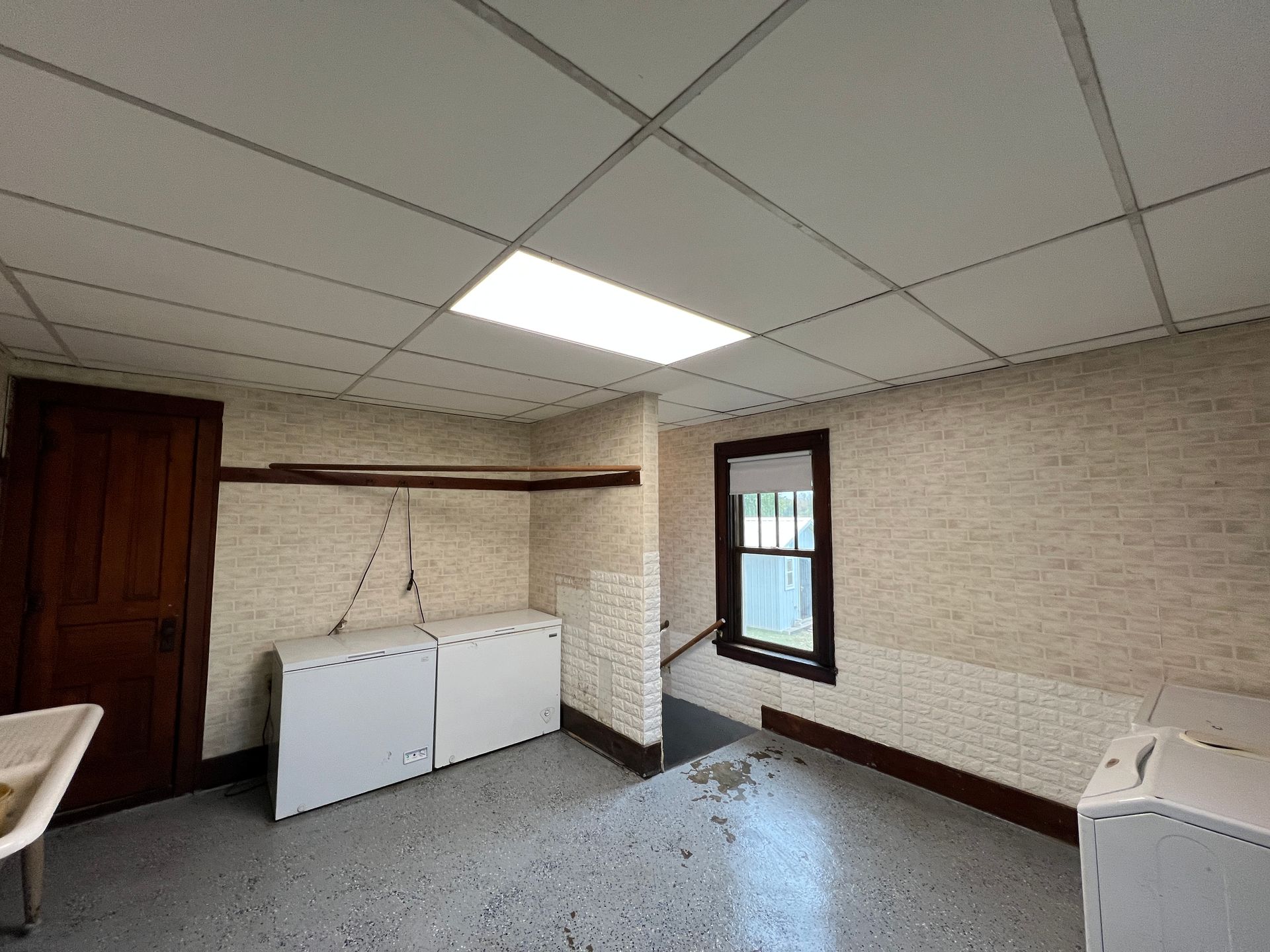 A room with a drop ceiling, two white chest freezers, a wooden door, a window, and stairs leading down to a dark area.