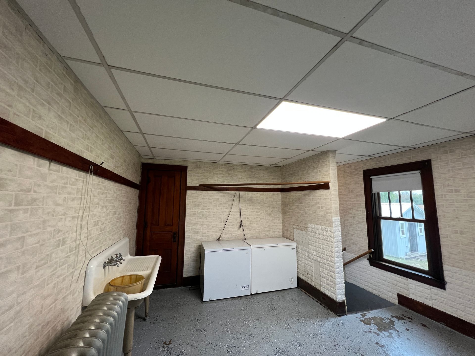 A room with light textured walls, a white drop ceiling, a vintage wall-mounted sink, two chest freezers, and a window.