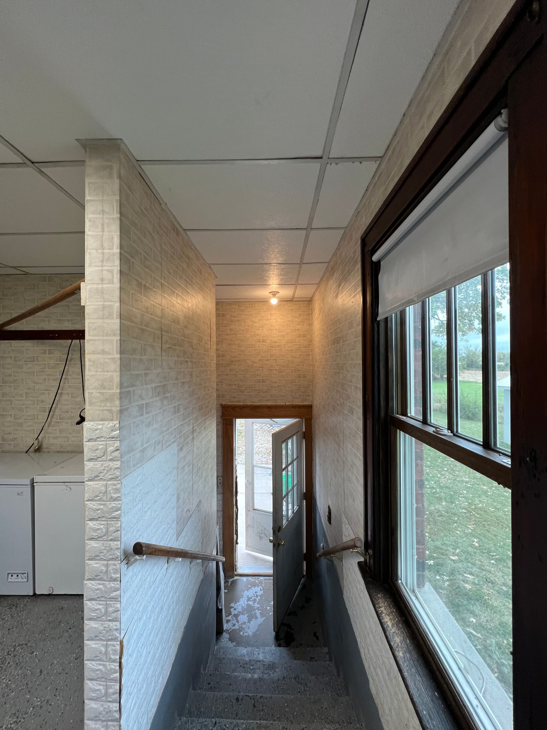A view looking down a stairwell with white brick-style walls, a window on the right, and a door at the bottom.