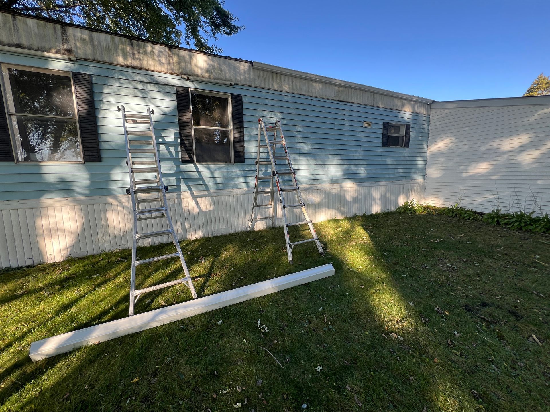 Two silver ladders lean against a light blue, horizontal-sided mobile home on a sunny day.