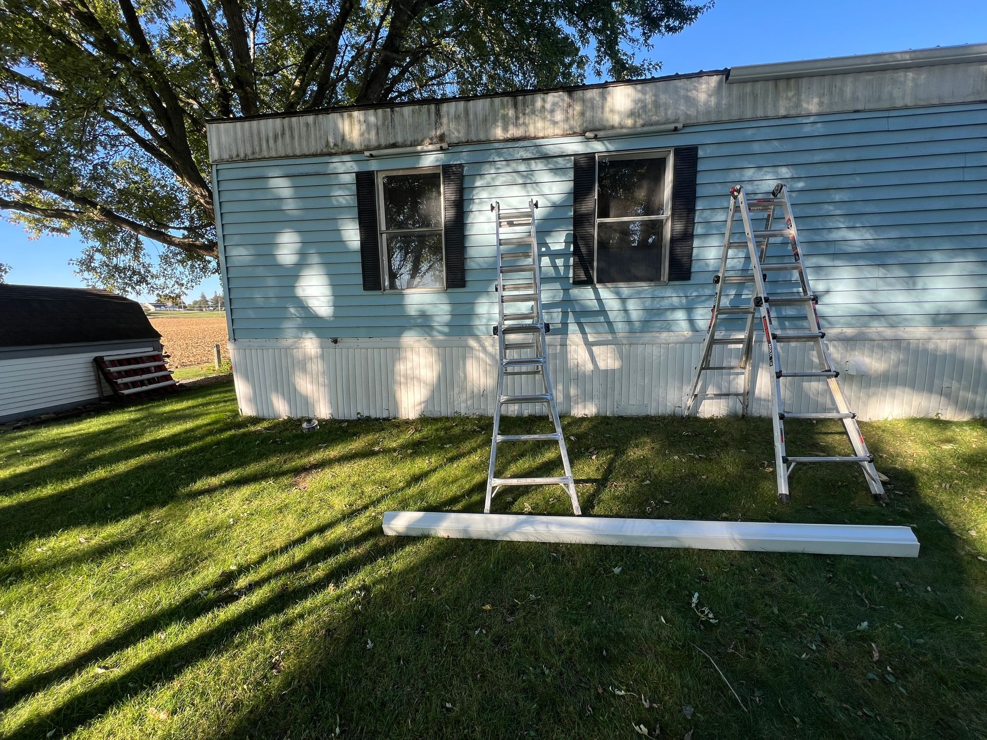 A light blue mobile home with two ladders in the yard and a long, white piece of trim lying on the grass in front.