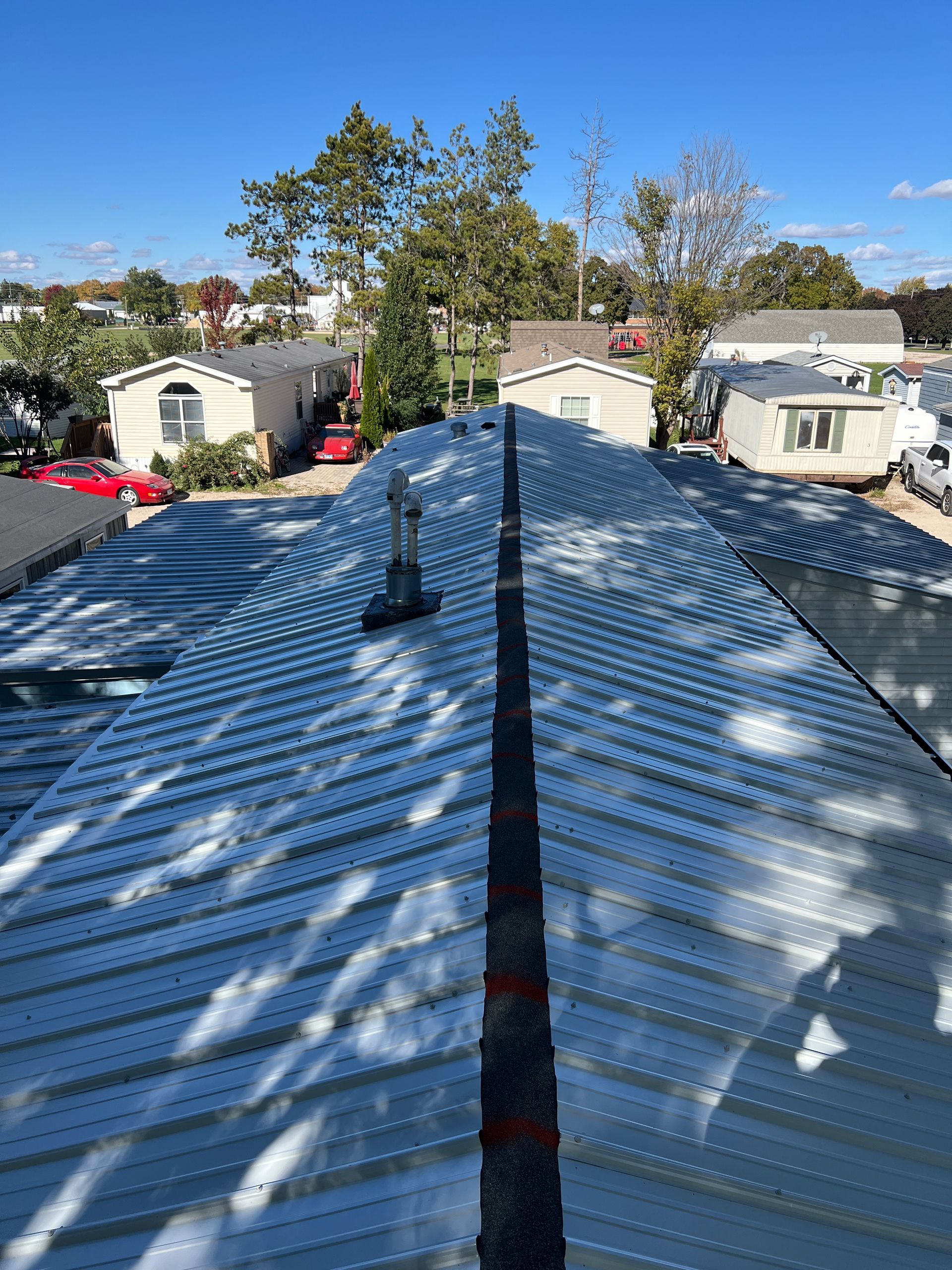 View of a residential metal roof with a central ridge cap, surrounded by other manufactured homes under a blue sky.