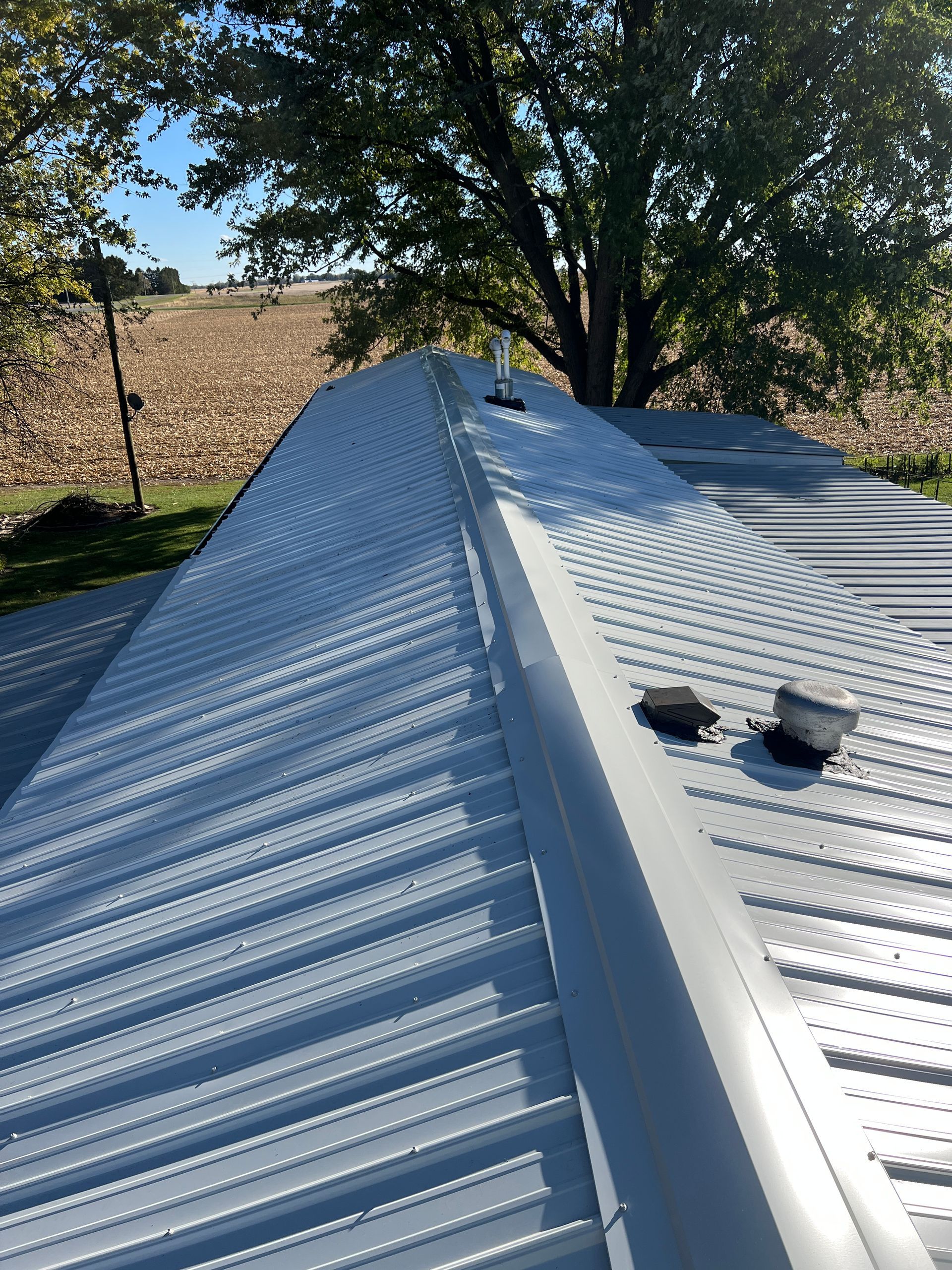 A metal roof ridge with a central vent, pipes, and a view of a field and tree in the background.