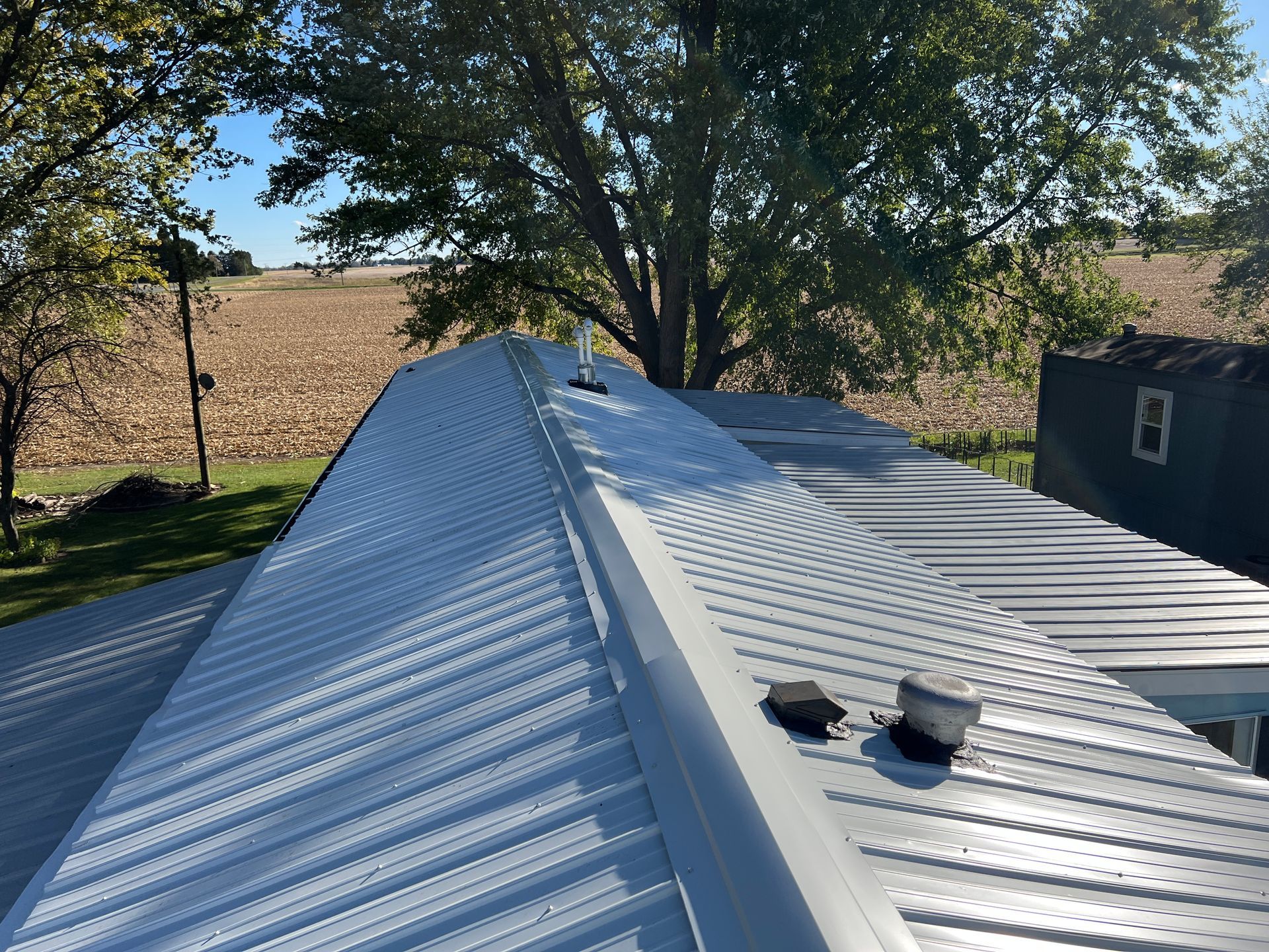 A high-angle view of a white corrugated metal roof on a sunny day, with trees and a harvested field in the background.