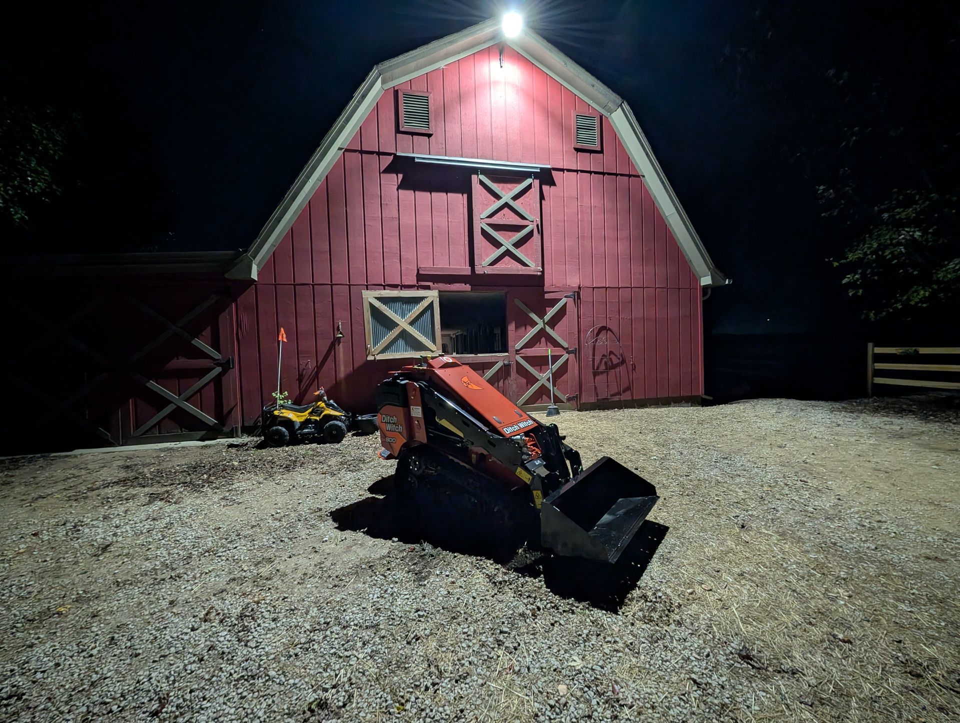 A red barn illuminated at night, with a skid steer loader parked on a gravel ground in the foreground.