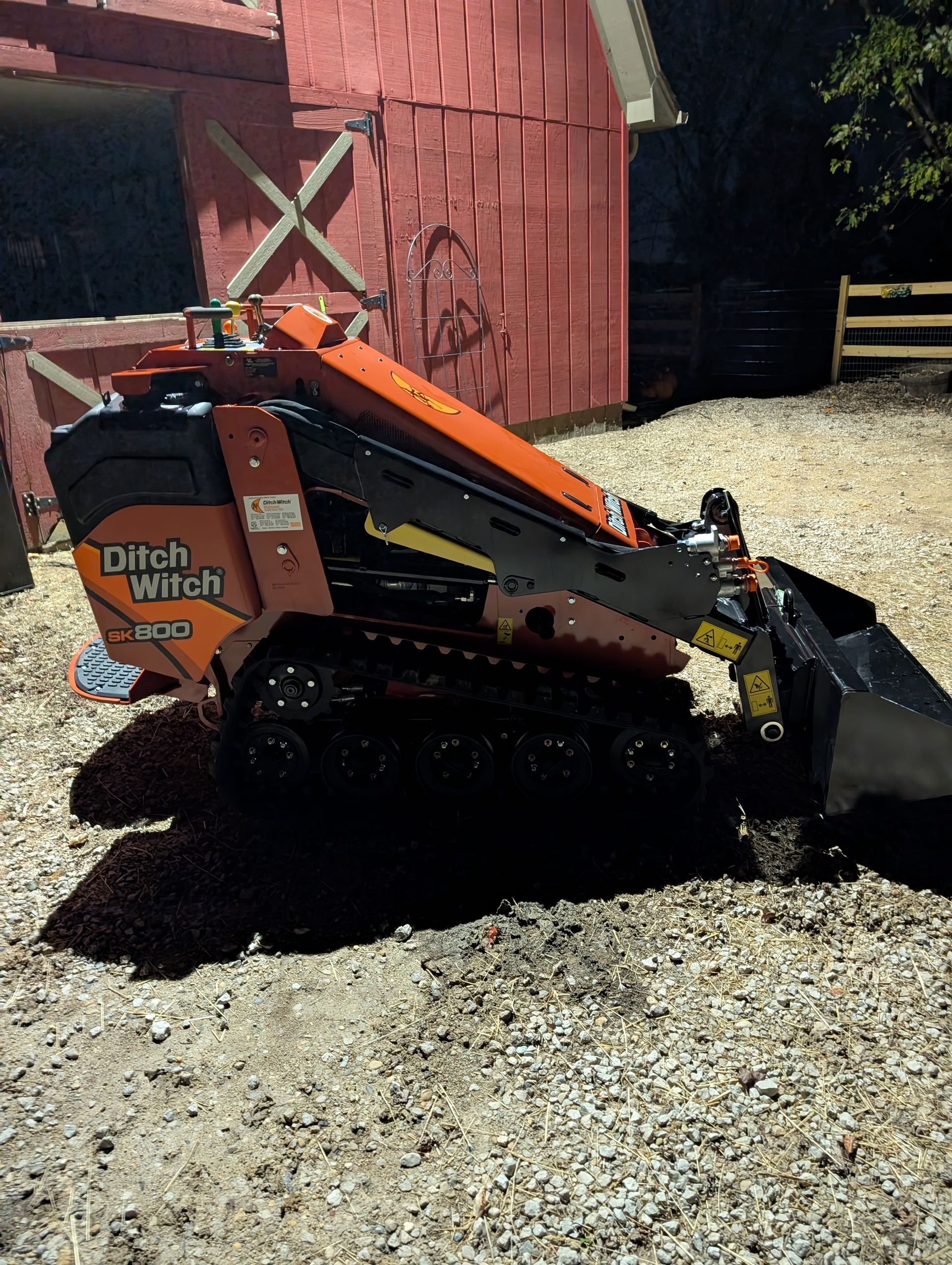 An orange Ditch Witch stand-on skid steer with tracks and a front bucket, parked on a gravel surface near a red building.