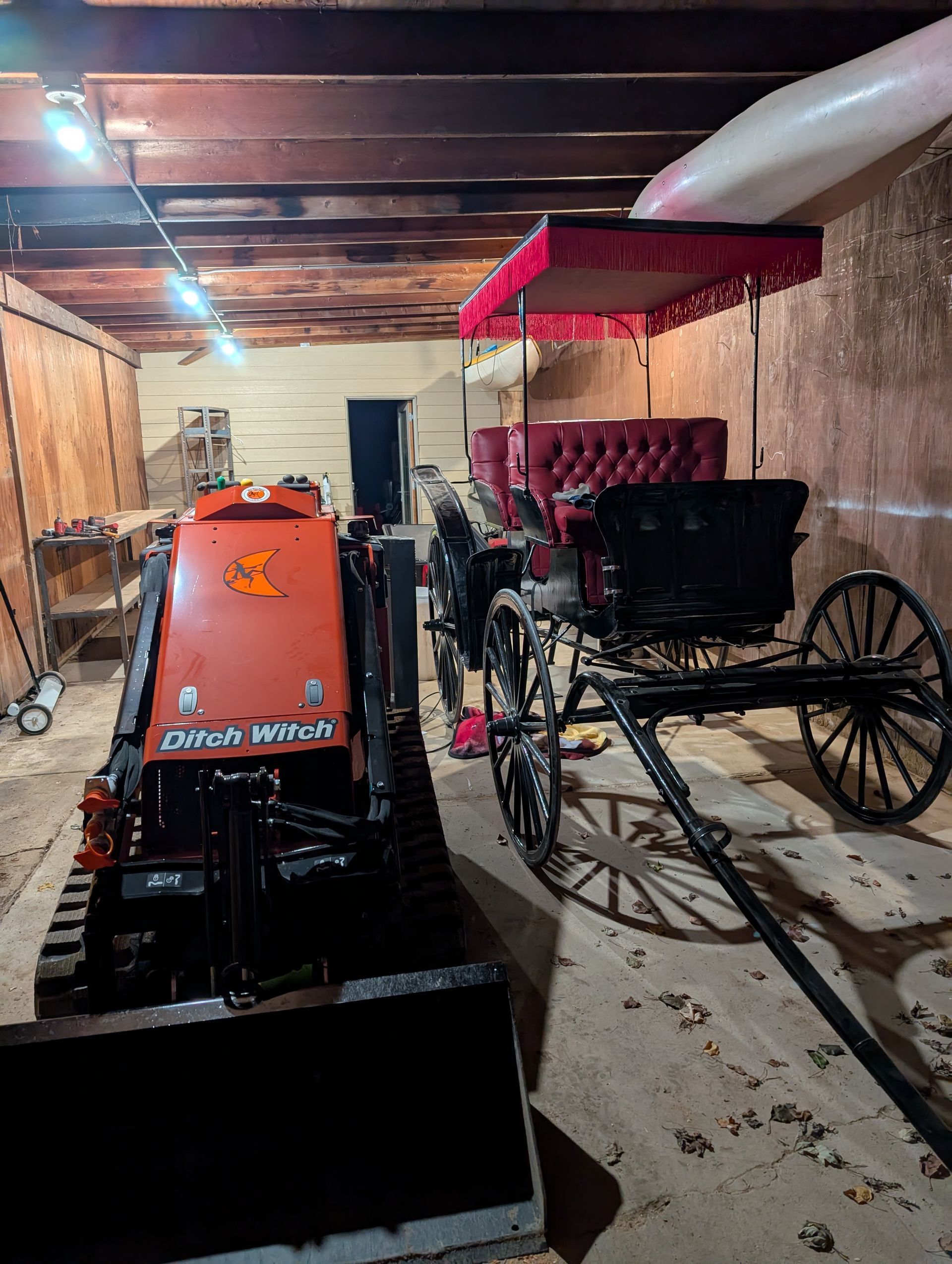 An orange tracked skid steer sits next to a black horse-drawn carriage with a red canopy inside a rustic wooden barn.