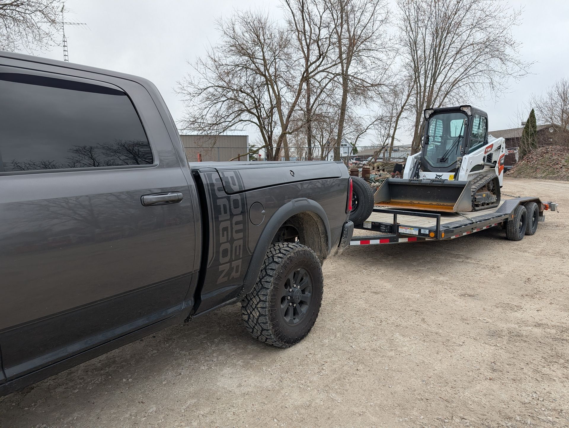A grey pickup truck towing a flatbed trailer with a white skid steer loader on a gravel lot.