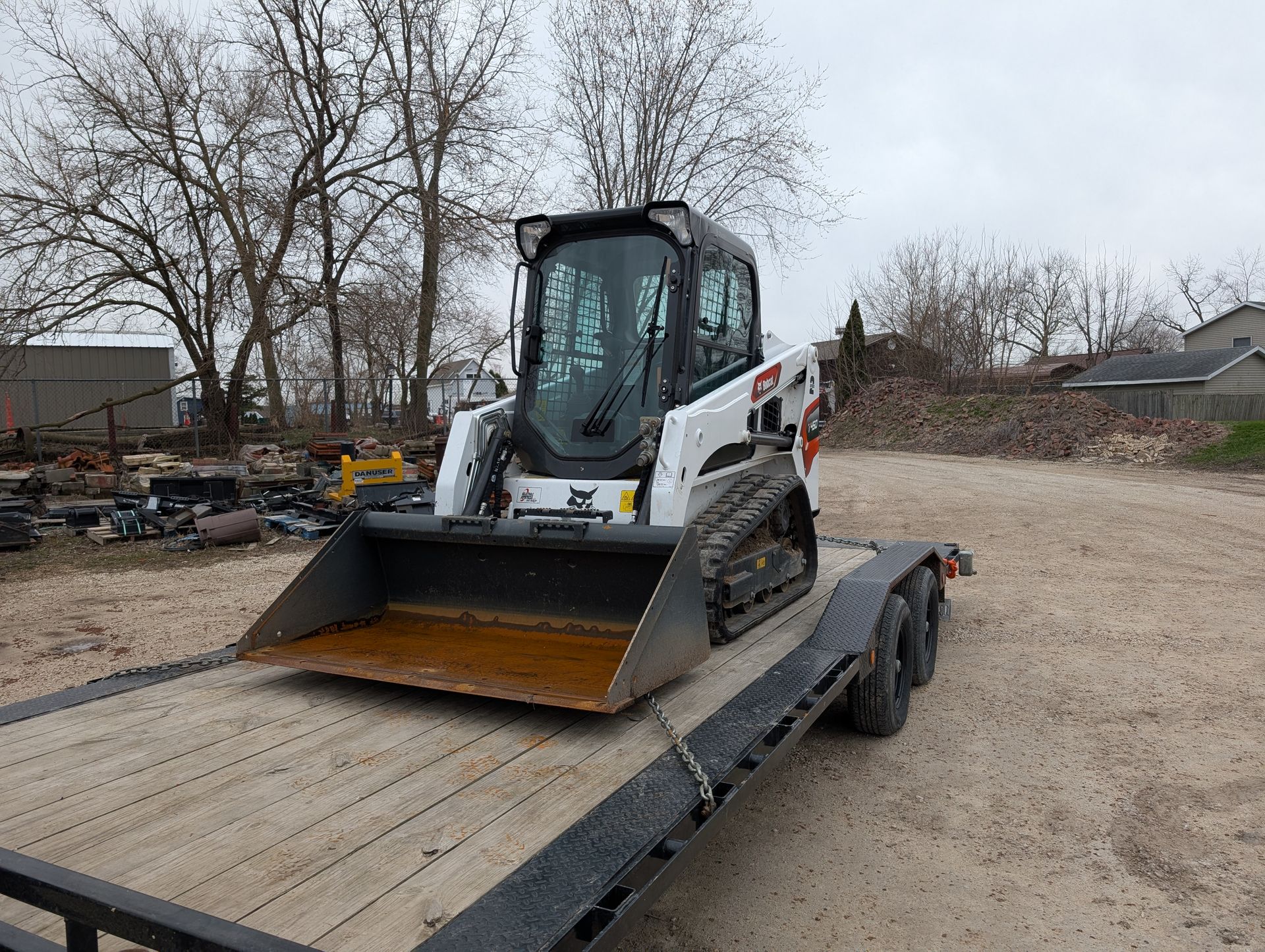 A white Bobcat compact track loader sits parked on a flatbed trailer in an outdoor, gravel-covered lot.