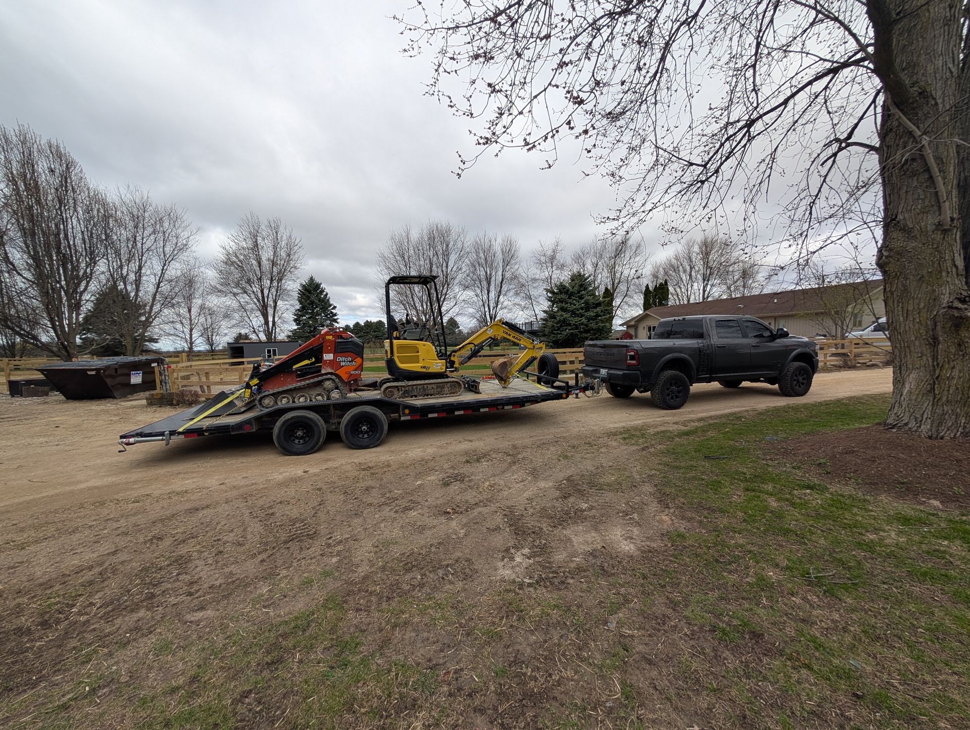 A black pickup truck tows a flatbed trailer carrying an orange skid steer and a yellow mini excavator on a gravel lot.