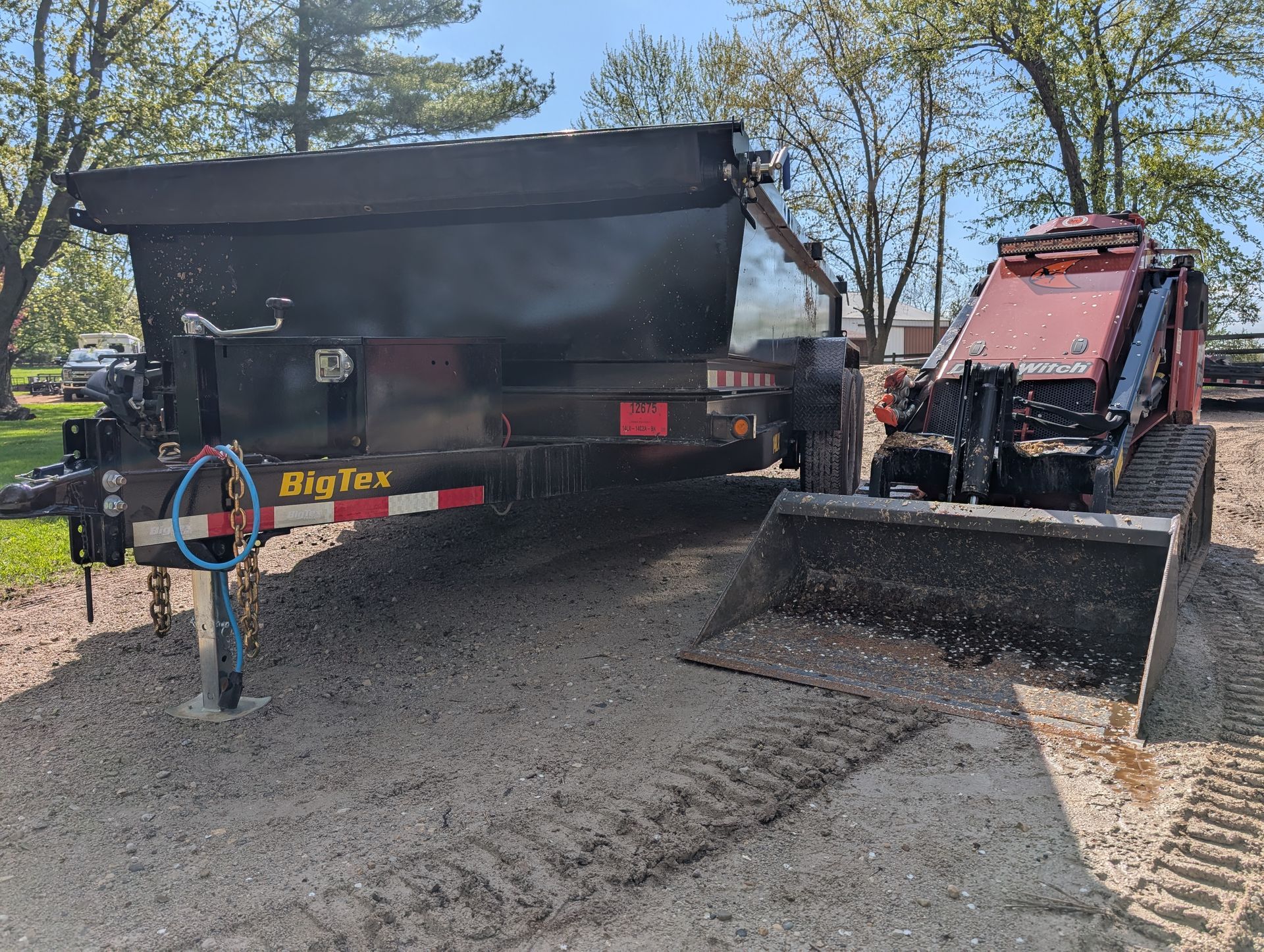 A red tracked skid steer loader parked next to a black Big Tex dump trailer on a gravel lot under a bright, sunny sky.