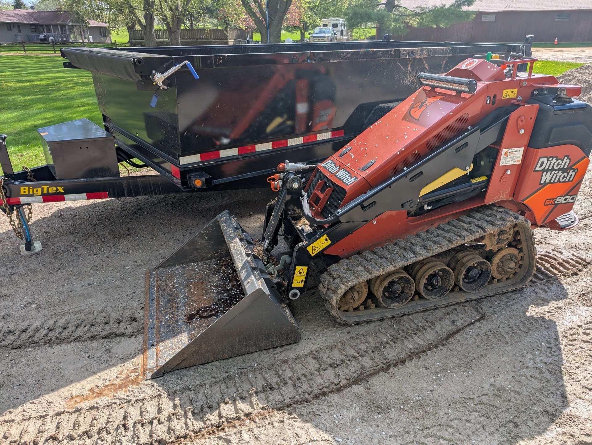 An orange Ditch Witch skid steer with a front bucket parked next to a black dump trailer on a gravel lot.
