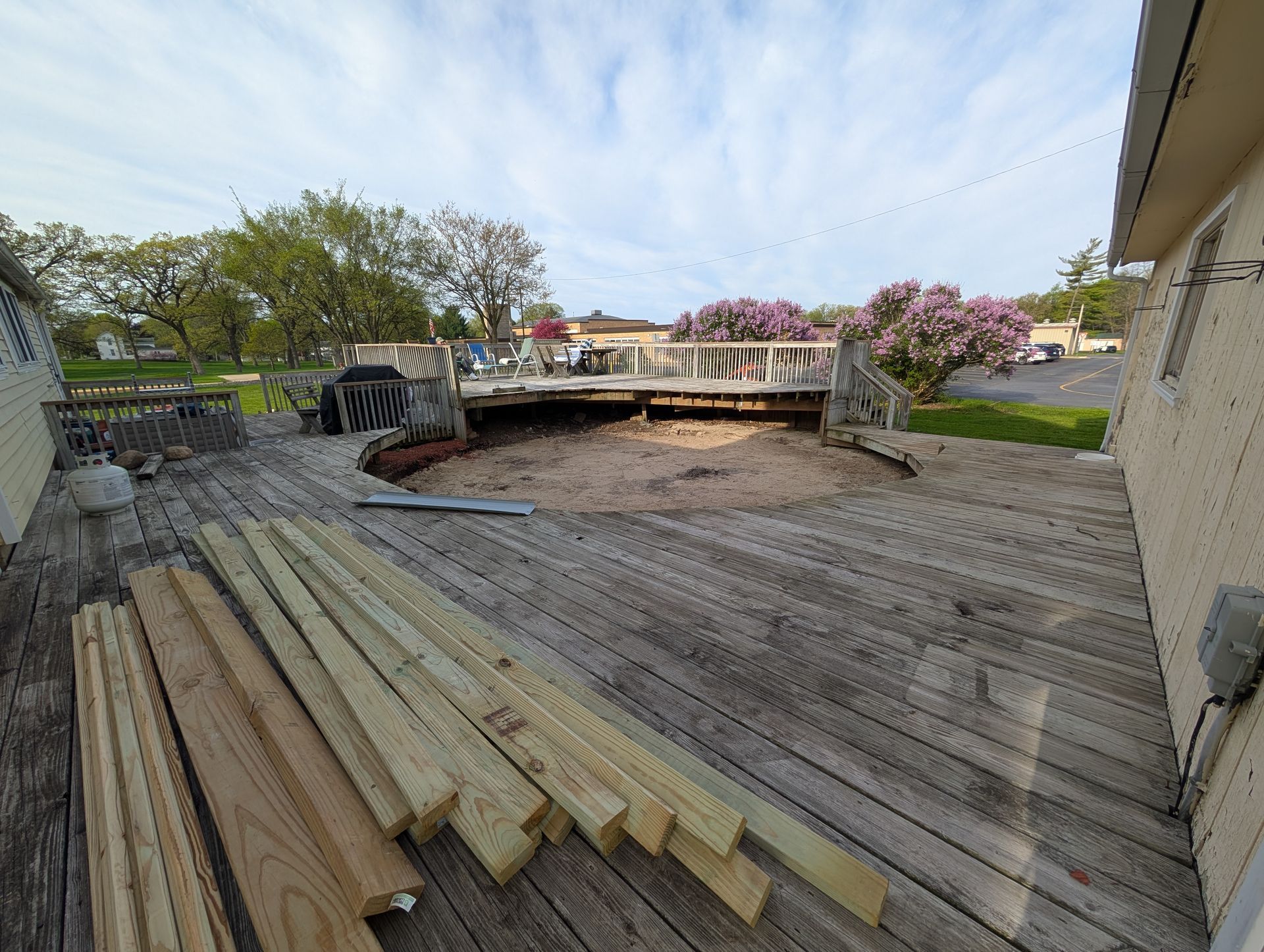 A wooden deck with a large circular hole in the center, surrounded by stacked lumber for repairs outdoors.