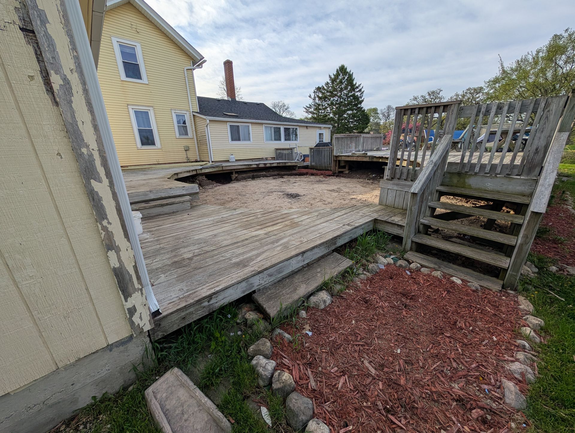 A yellow house with a large, partially collapsed wooden deck and stone landscaping in the backyard.