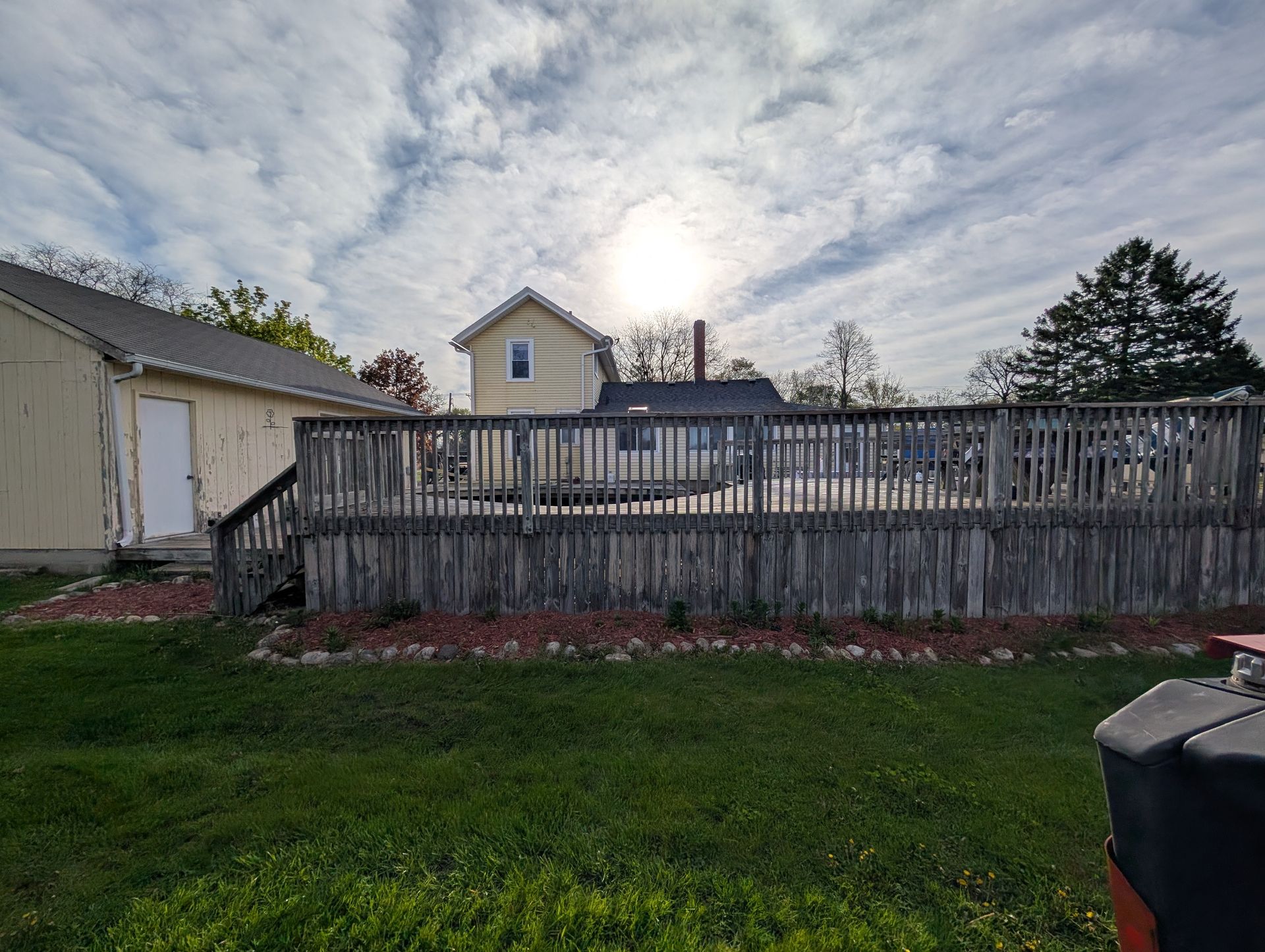 A yellow house with a large wooden deck in the backyard under a cloudy, bright sky.