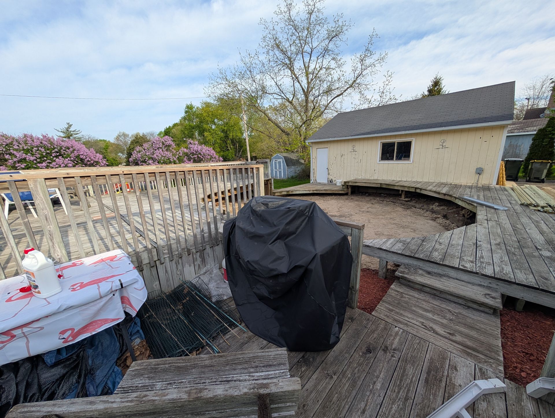 A weathered wooden deck wraps around an empty above-ground pool area next to a light-colored garage on a sunny day.