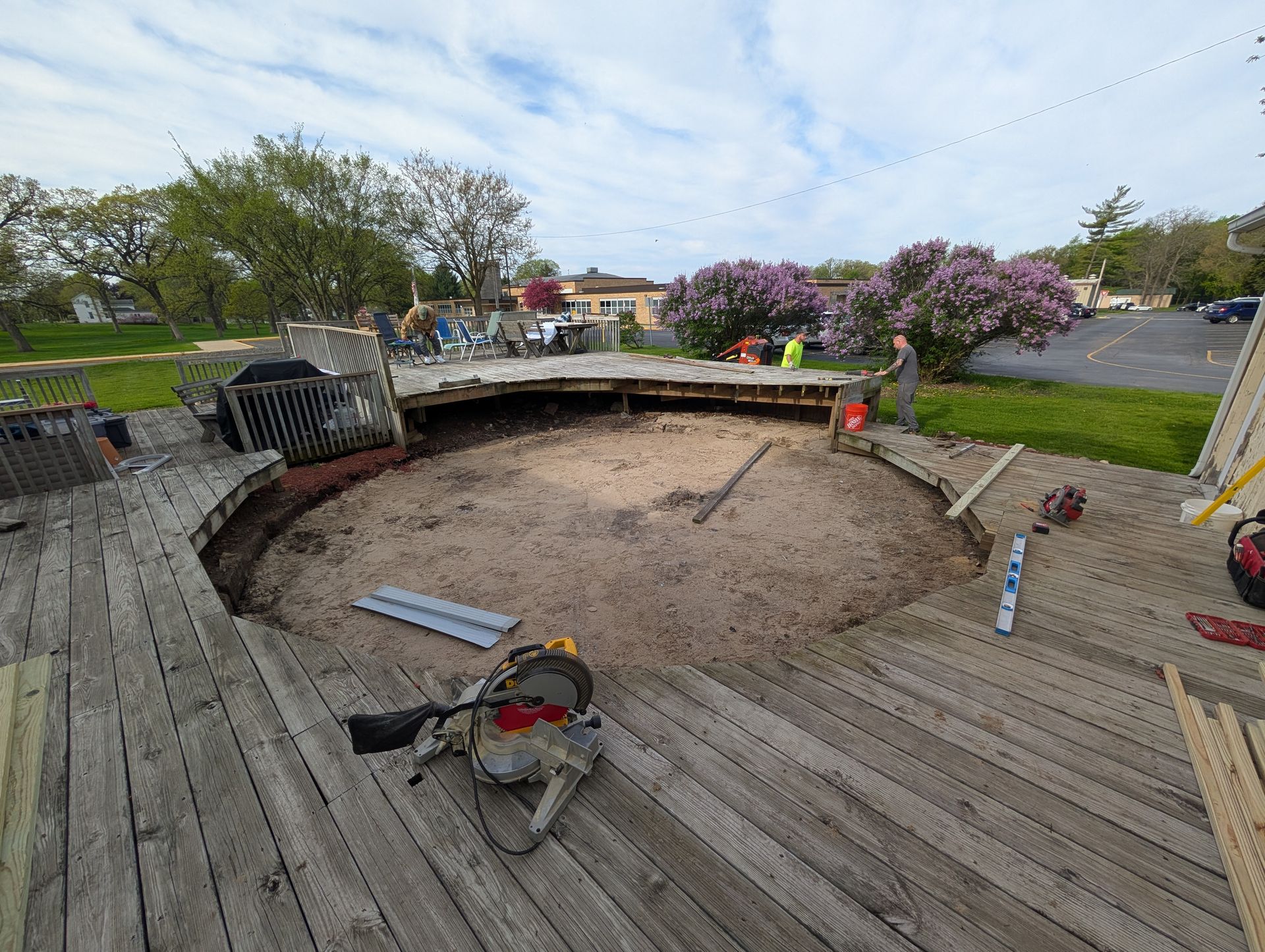 Workers renovate a circular backyard deck surrounding a sand-filled pit under a cloudy sky.