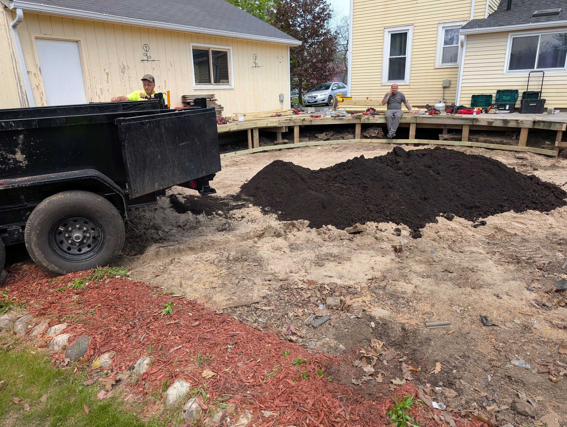 A person sits on a trailer filled with dark soil in a yard between two houses while another person stands nearby.
