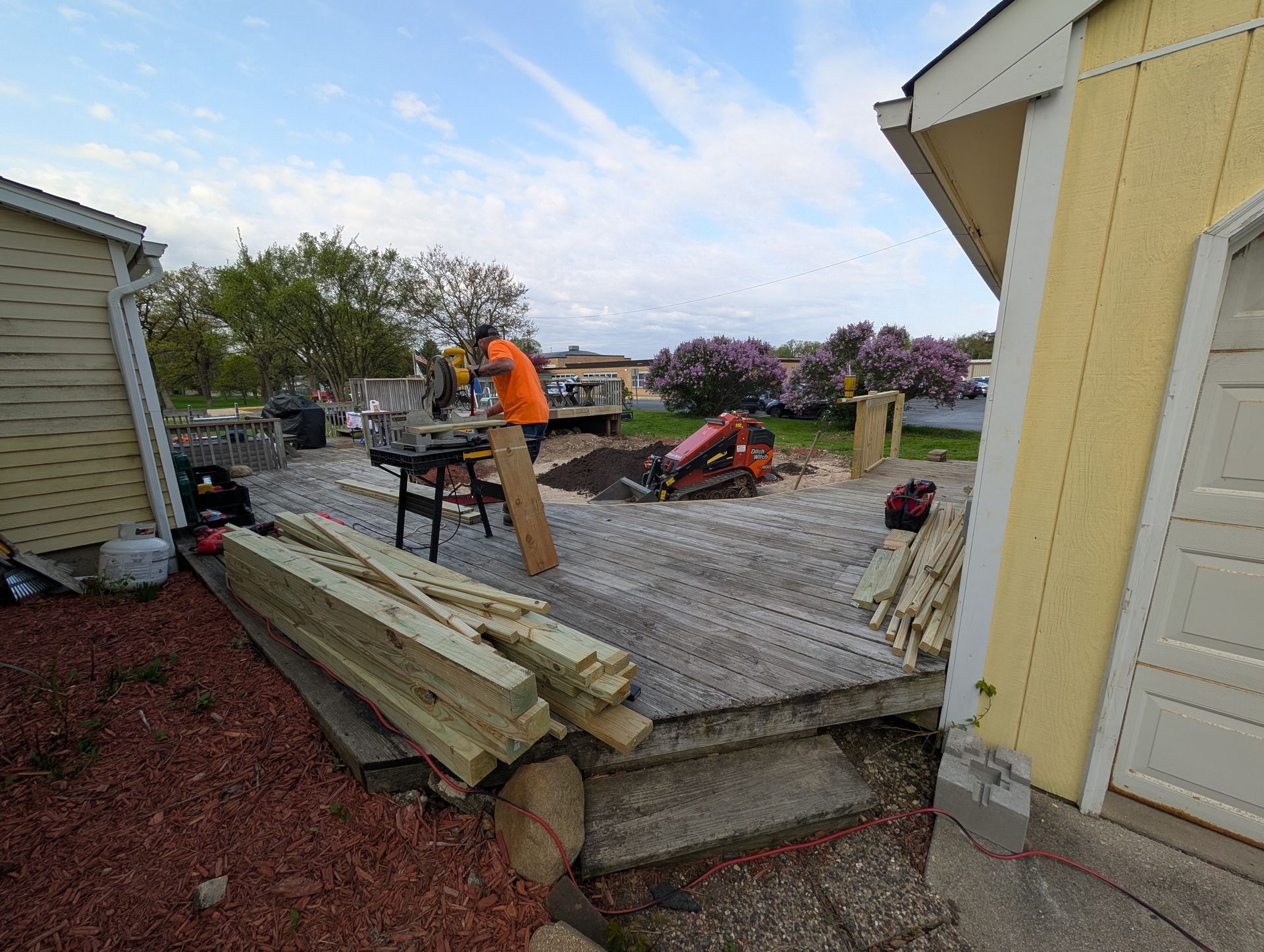 A person in an orange shirt works on a wooden deck project between two yellow-sided houses on a sunny day.