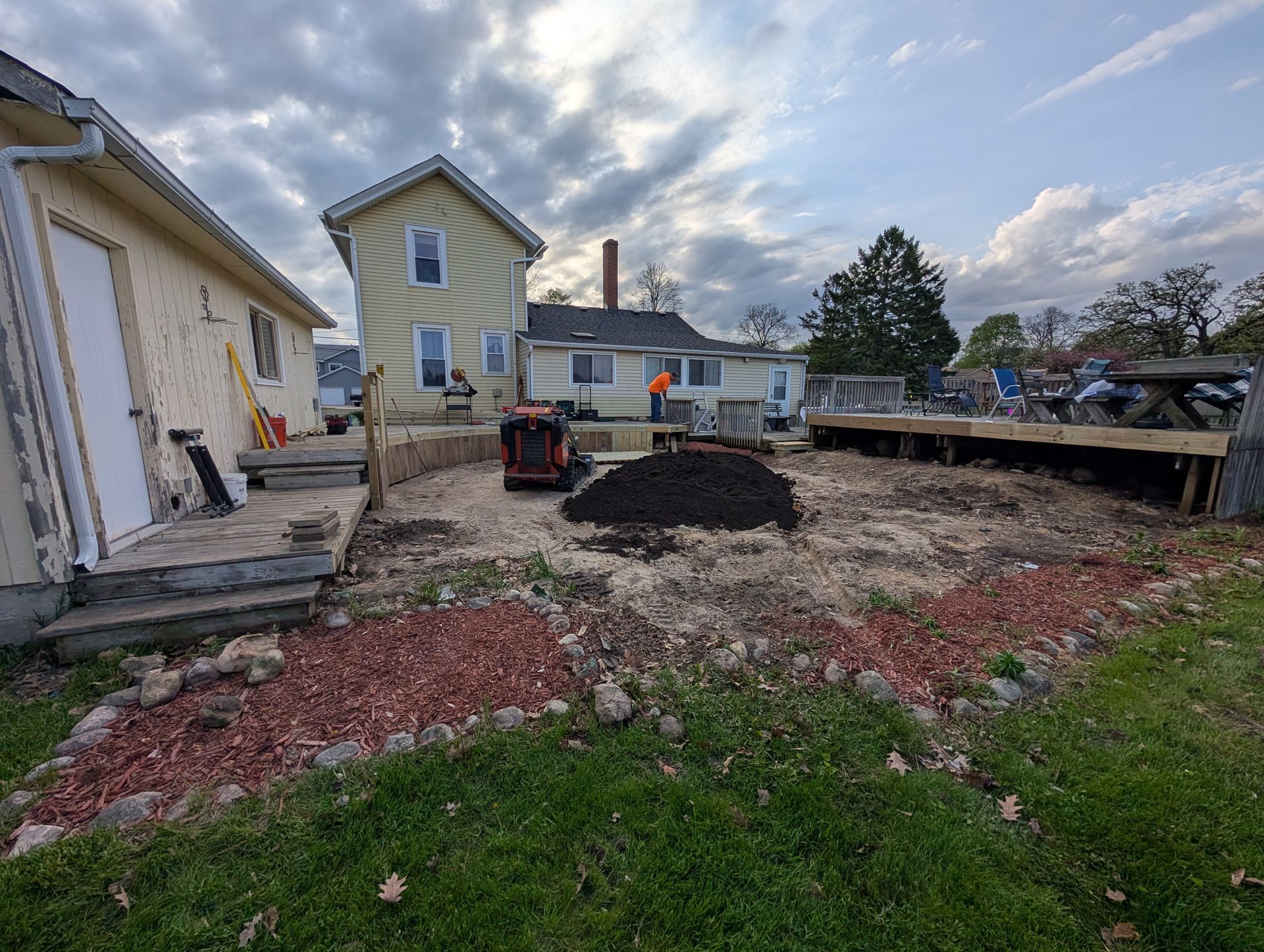 A backyard construction site featuring a partially built deck, a soil pile, a small loader, and yellow siding houses.
