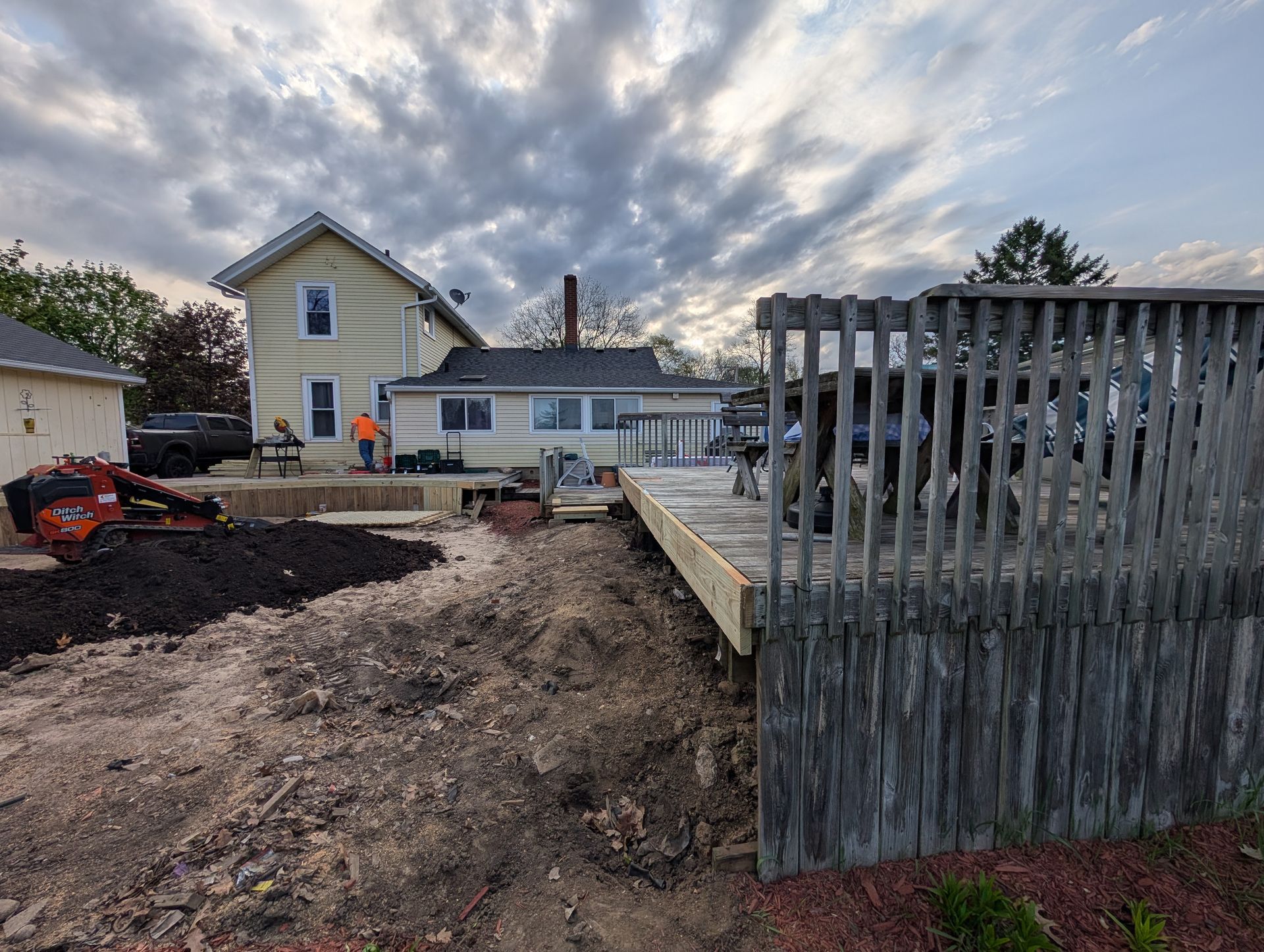 A construction crew works on a house deck exterior in a yard with a large pile of dark soil and an existing wood deck.