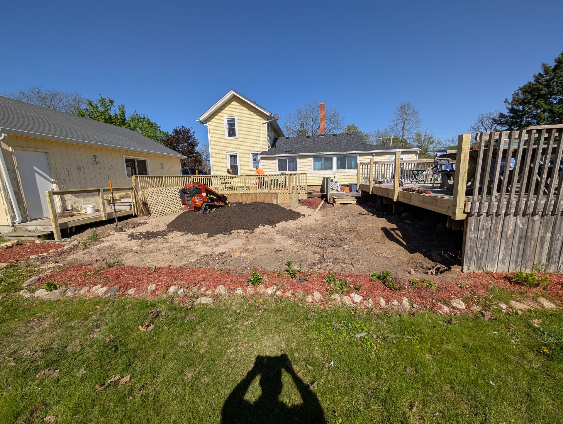 A person uses a rototiller in a backyard between two wooden decks and a house on a sunny day.