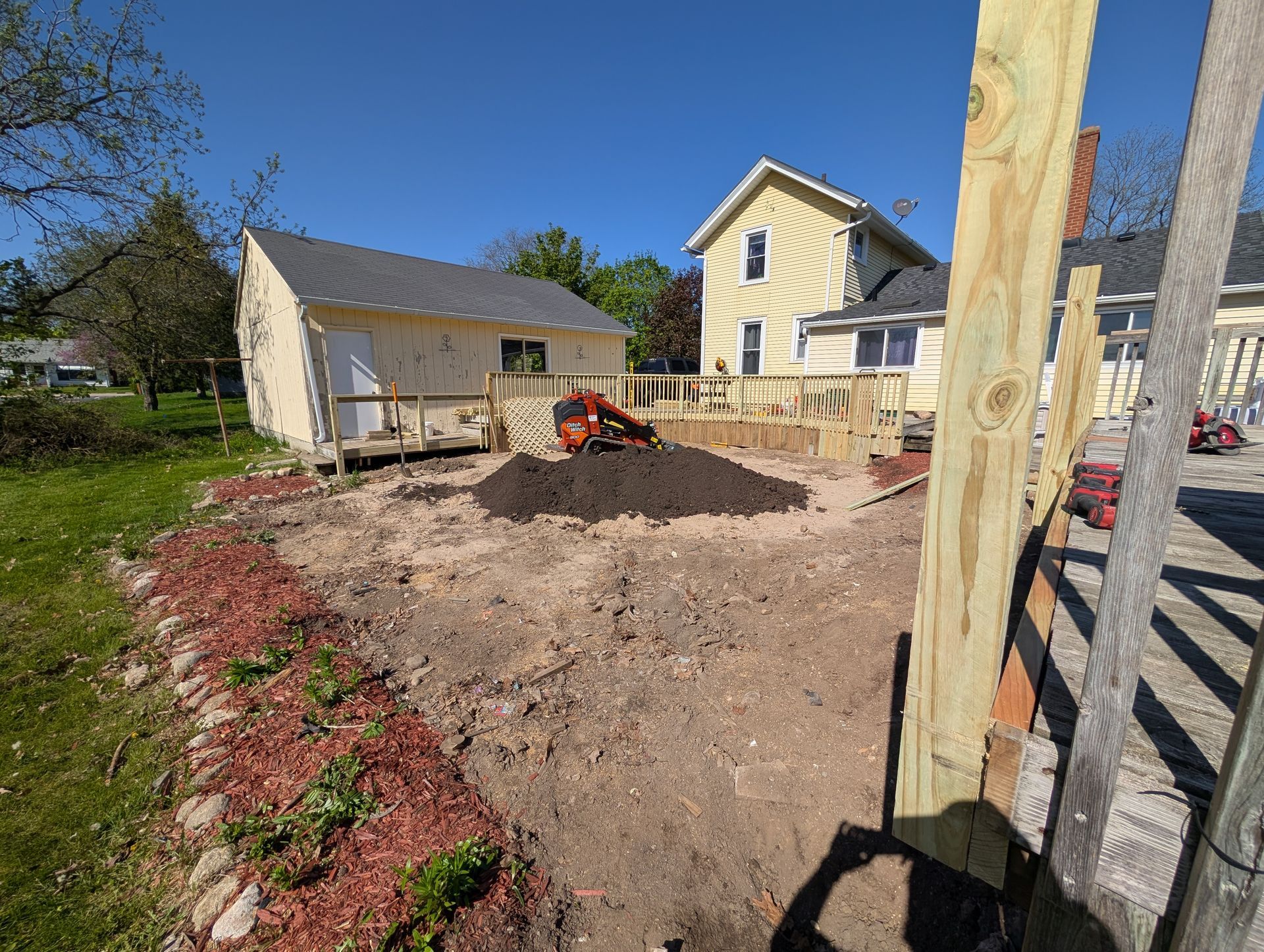 A construction site showing an excavated dirt yard, a small red tiller, and wooden framing near a house and a garage.