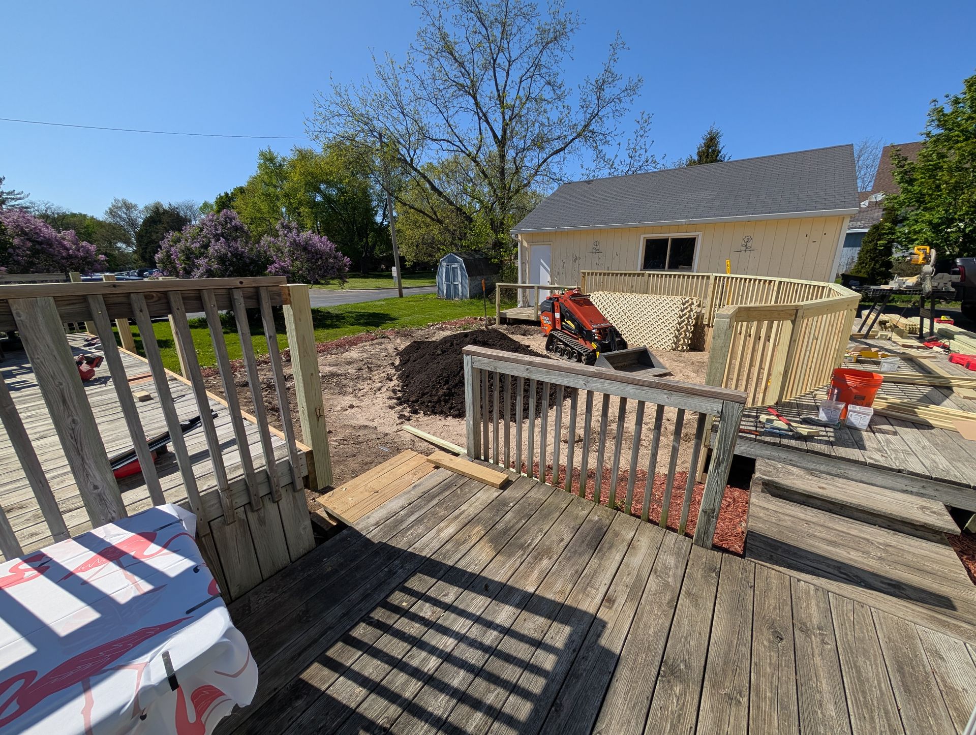 A wooden deck under construction with exposed soil, a small skid steer, and stacks of lumber in a sunny backyard.