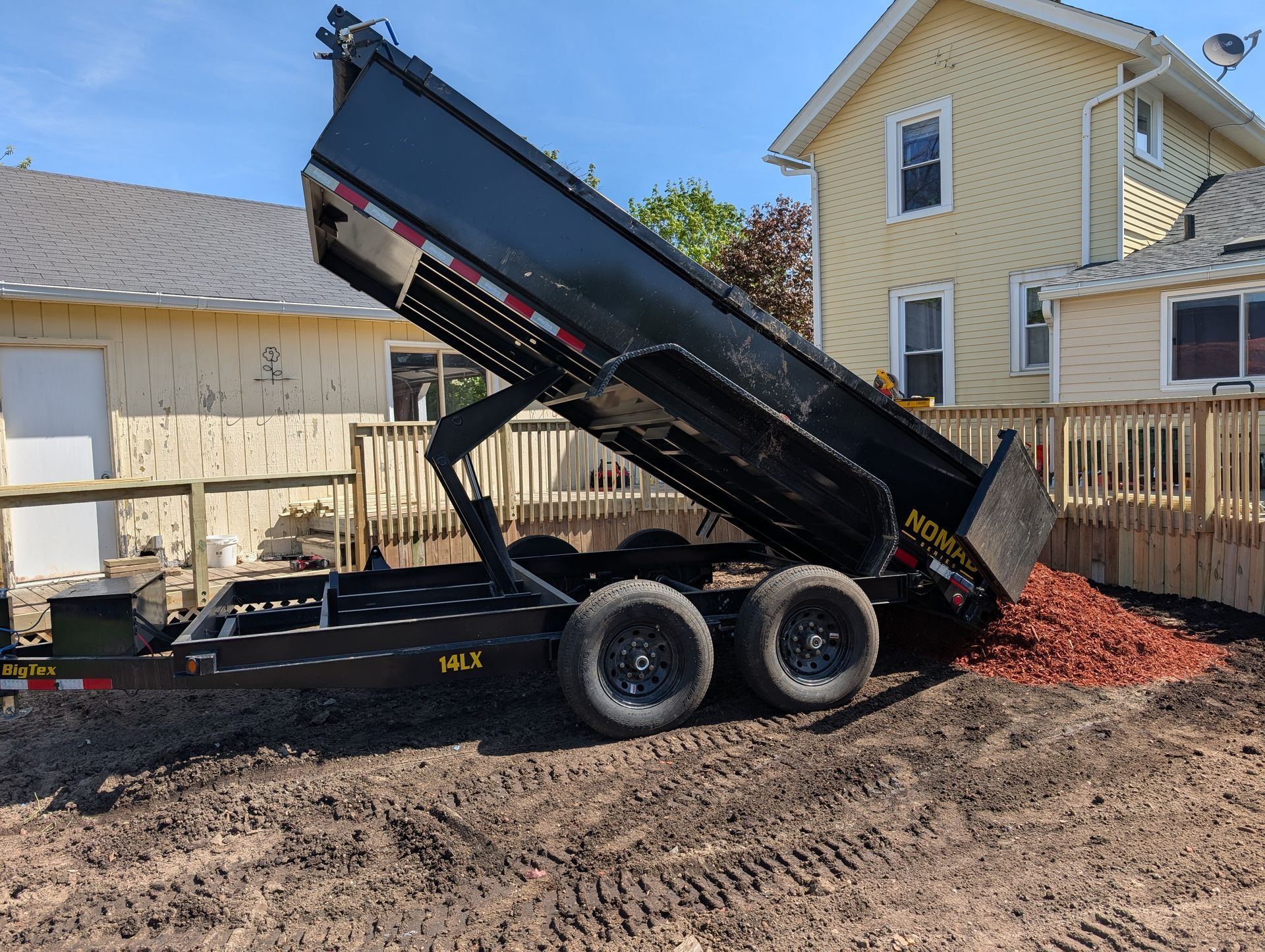 A black dump trailer raised to deposit a pile of reddish mulch on a residential lawn next to a yellow house.