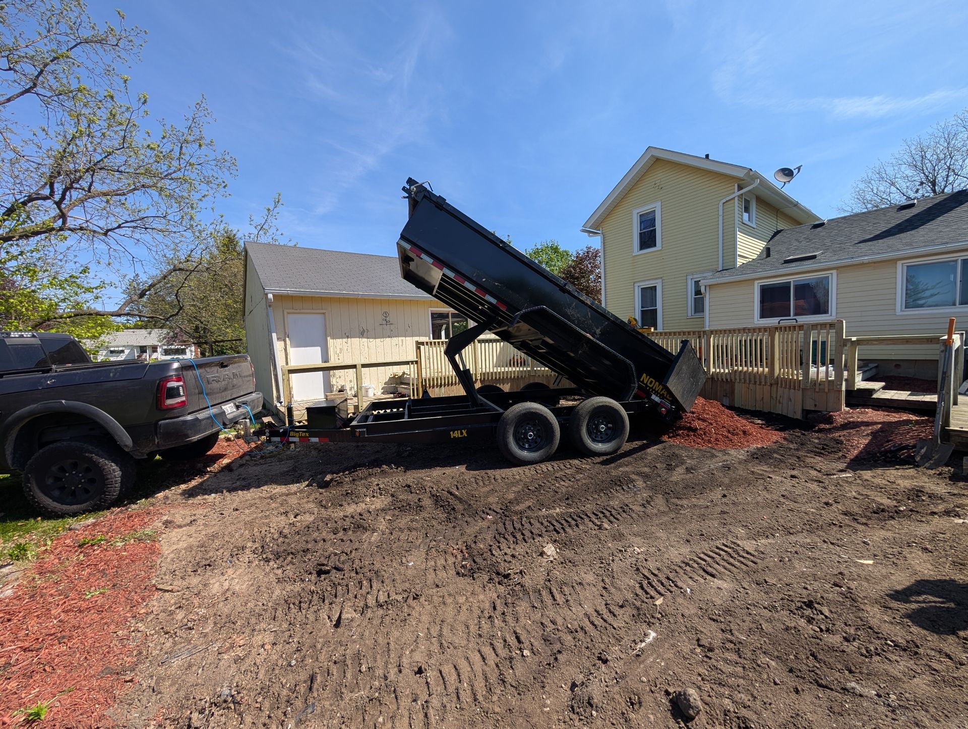 A dump trailer raised to unload red mulch next to a house and garage on a sunny day.