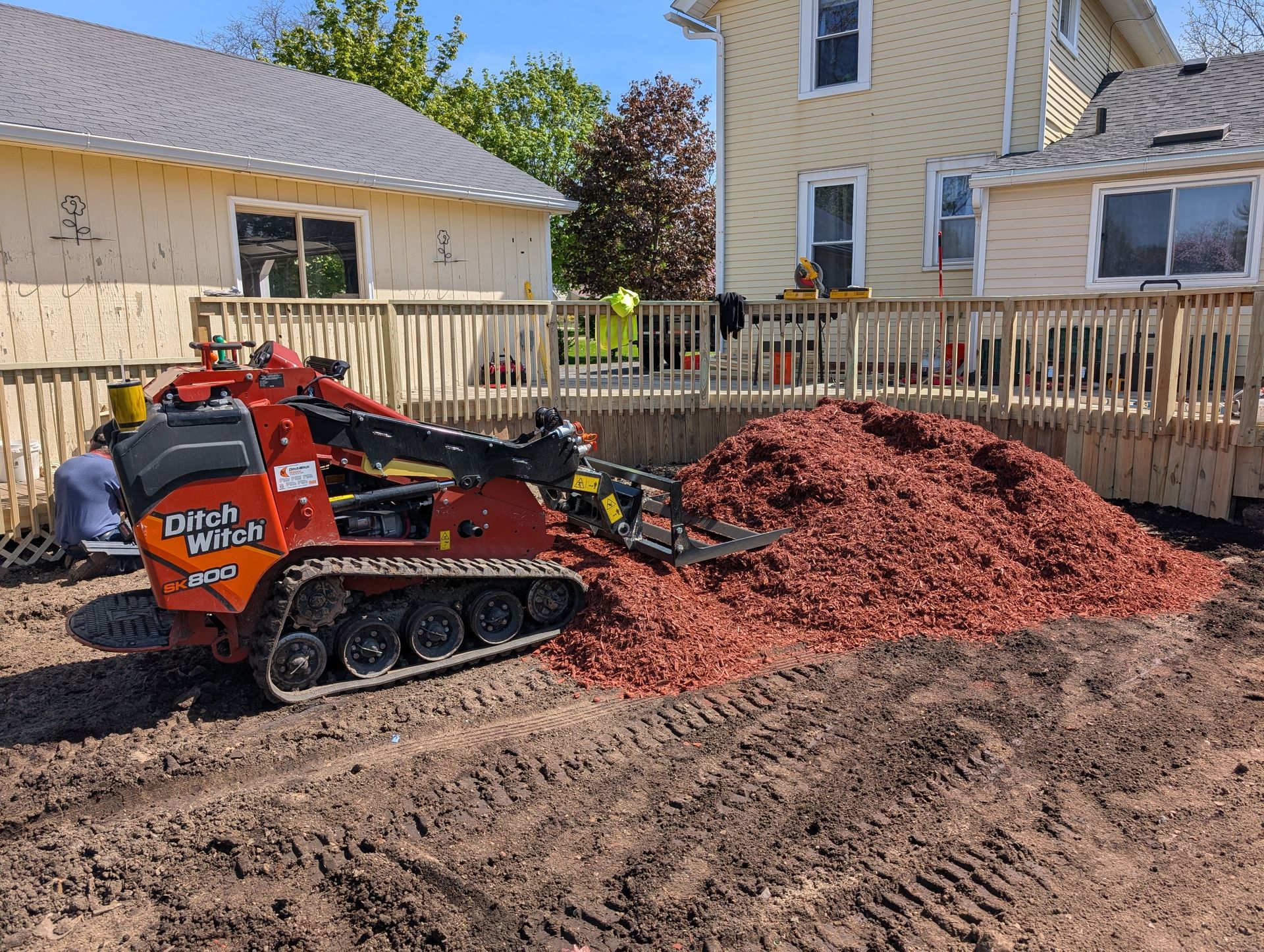 An orange Ditch Witch skid steer loader sits next to a large pile of red mulch in front of a house and deck.