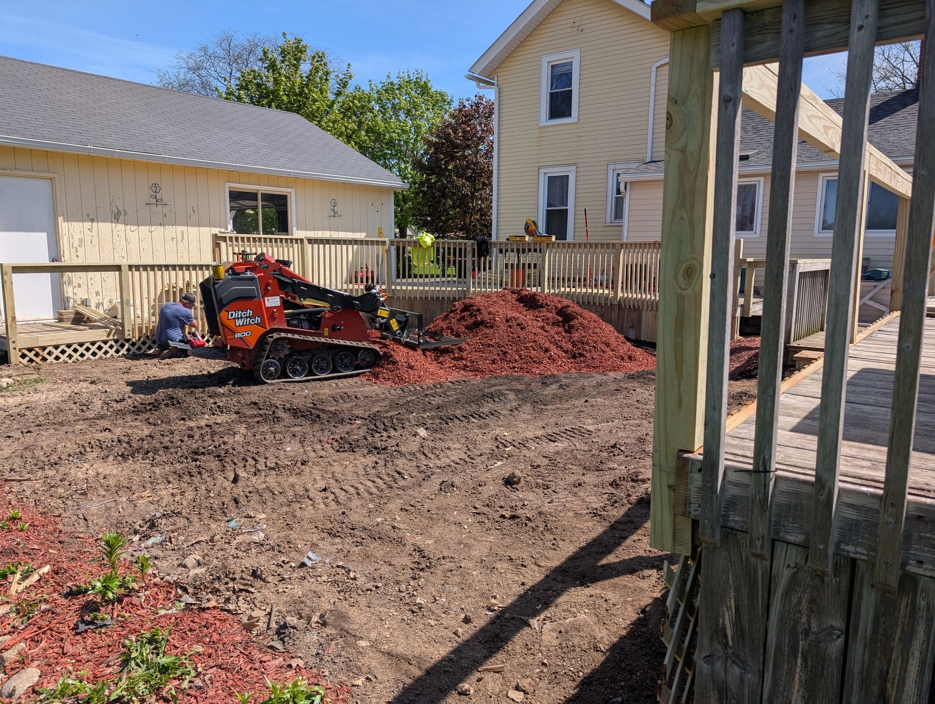 A worker operates a small orange tracked loader to spread red mulch in a residential yard near a house and wooden deck.