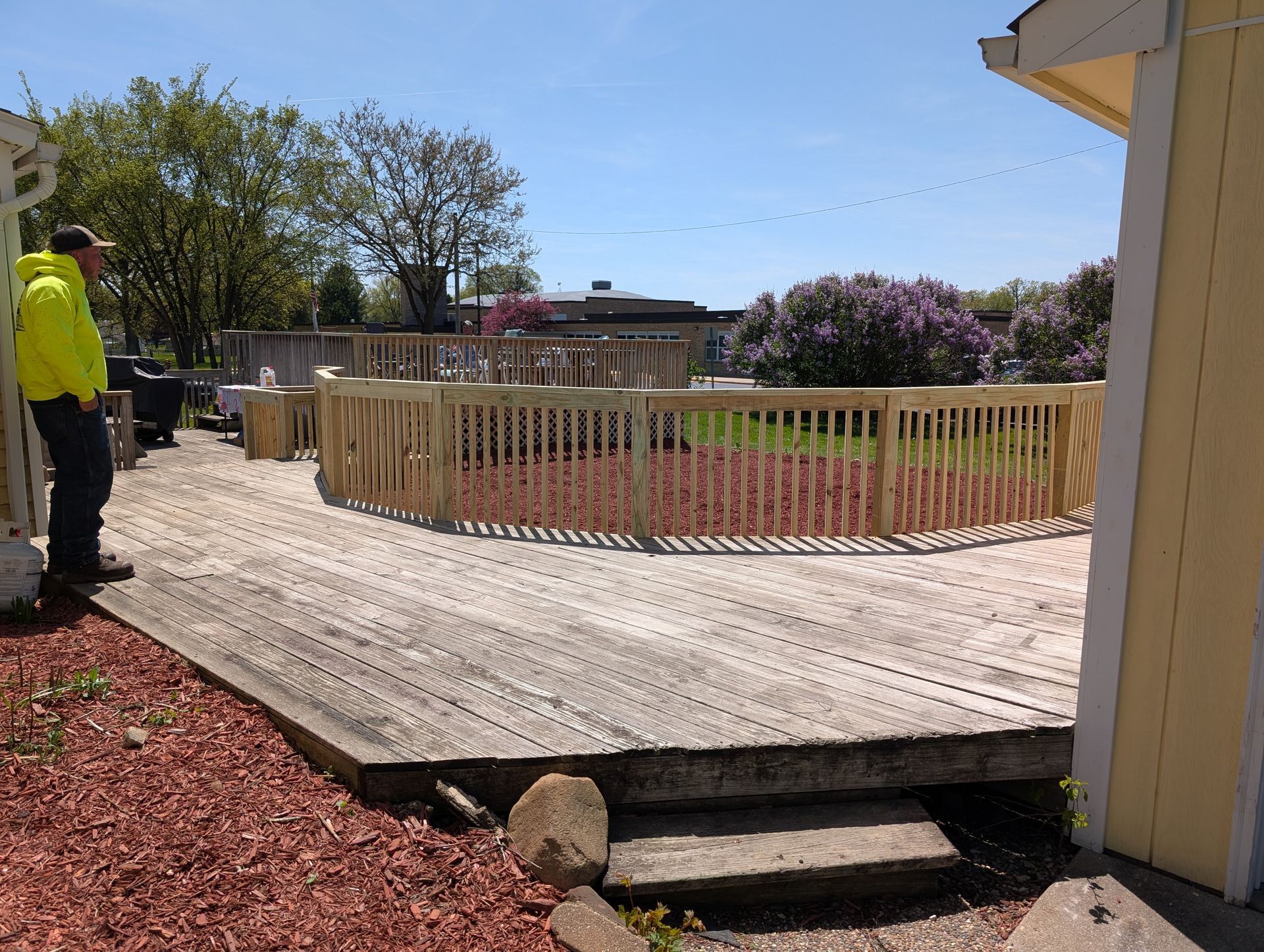 A person in a neon yellow jacket stands on a wooden deck next to a fenced-in garden area on a sunny day.