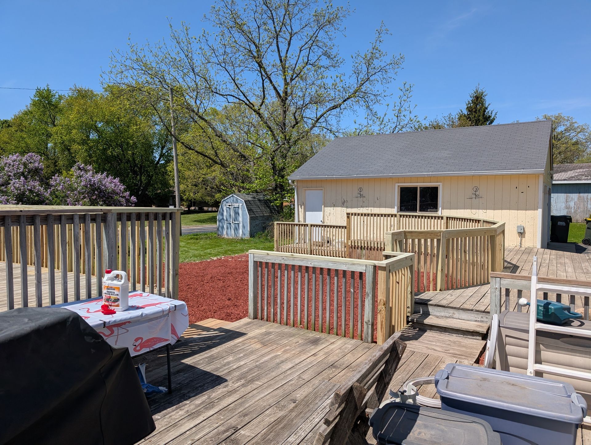 A wooden deck with a new wheelchair ramp, railing, and a barbecue grill on a sunny day in front of a light-colored garage.