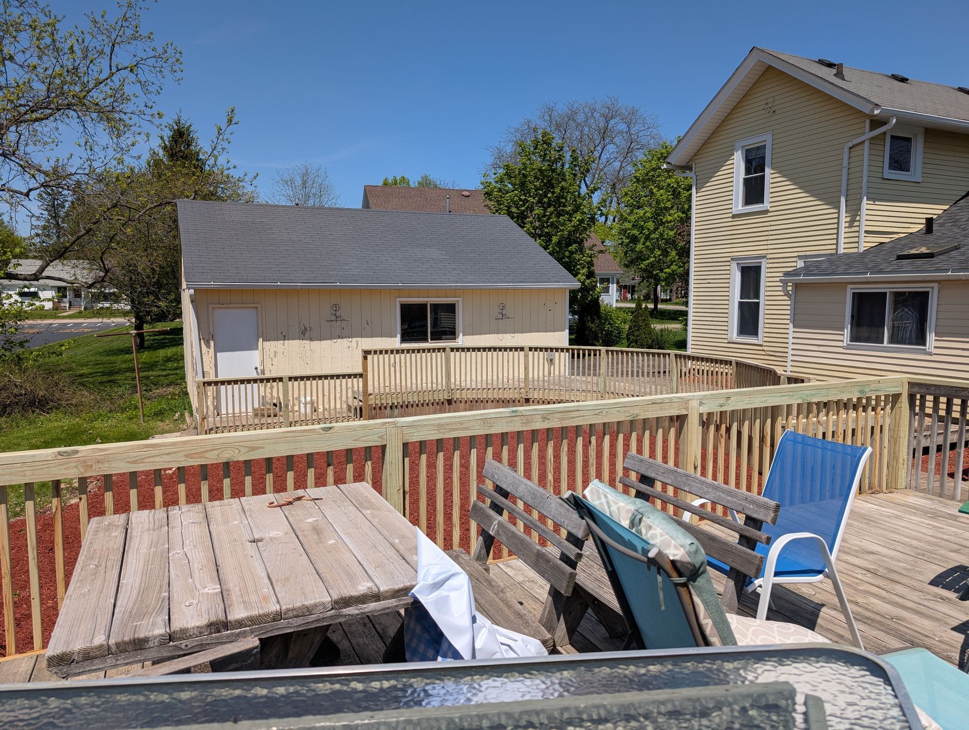 A wooden deck with a picnic table and chairs overlooks a backyard featuring a detached light-colored garage and a house.