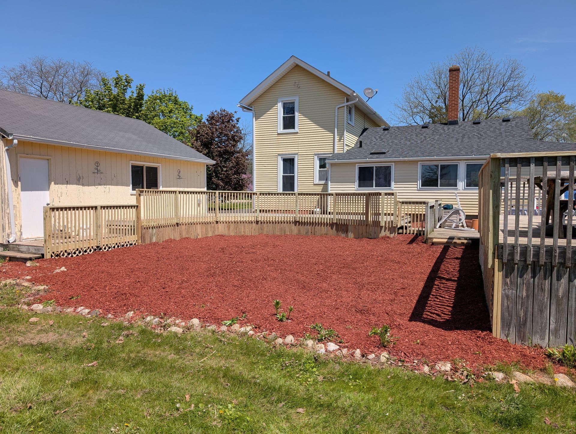 A yard with red mulch, wooden fencing, and two houses under a blue sky.