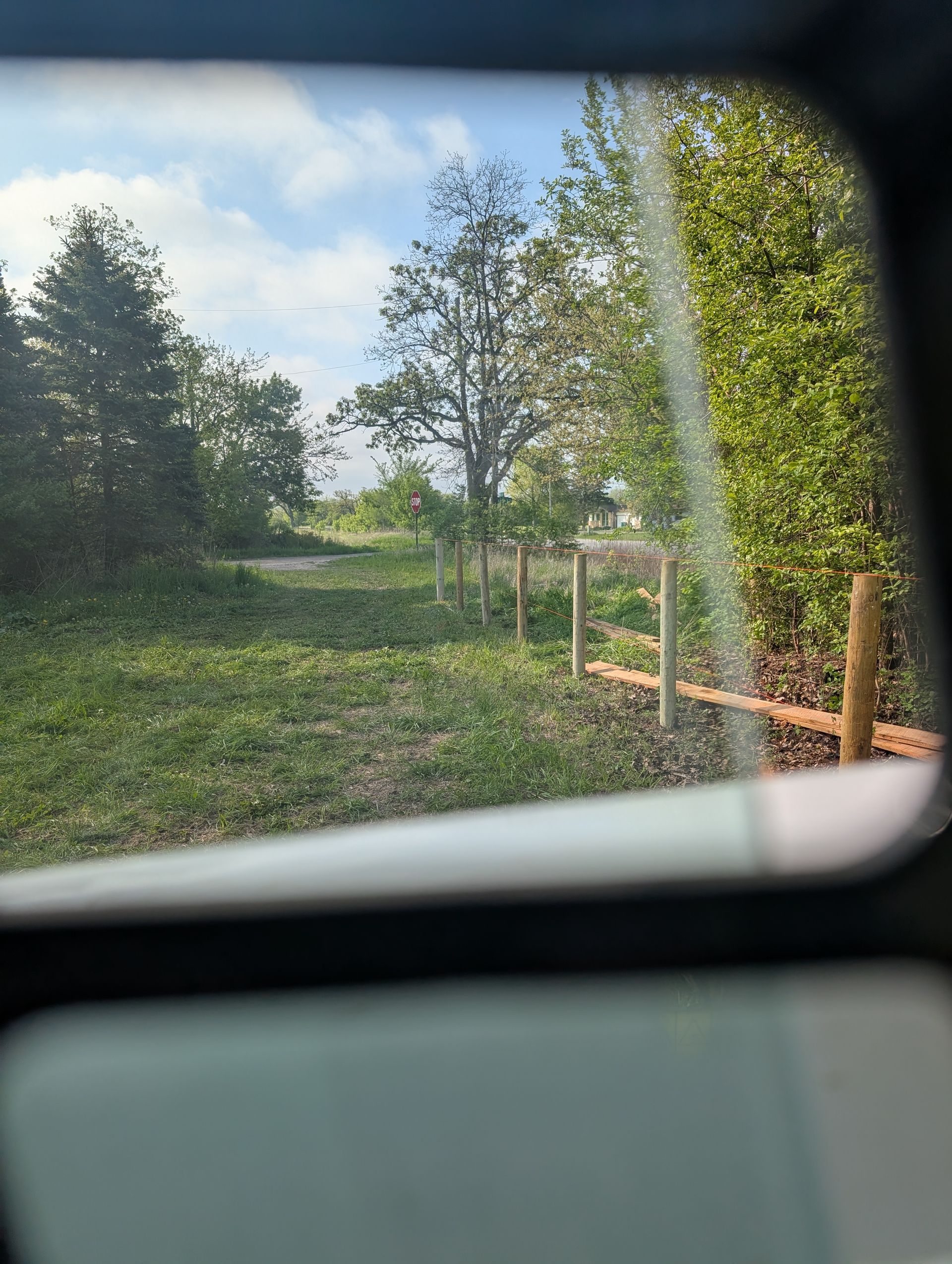 A rural landscape viewed through a car window, featuring green trees, a grassy field, and a wooden post-and-rail fence.