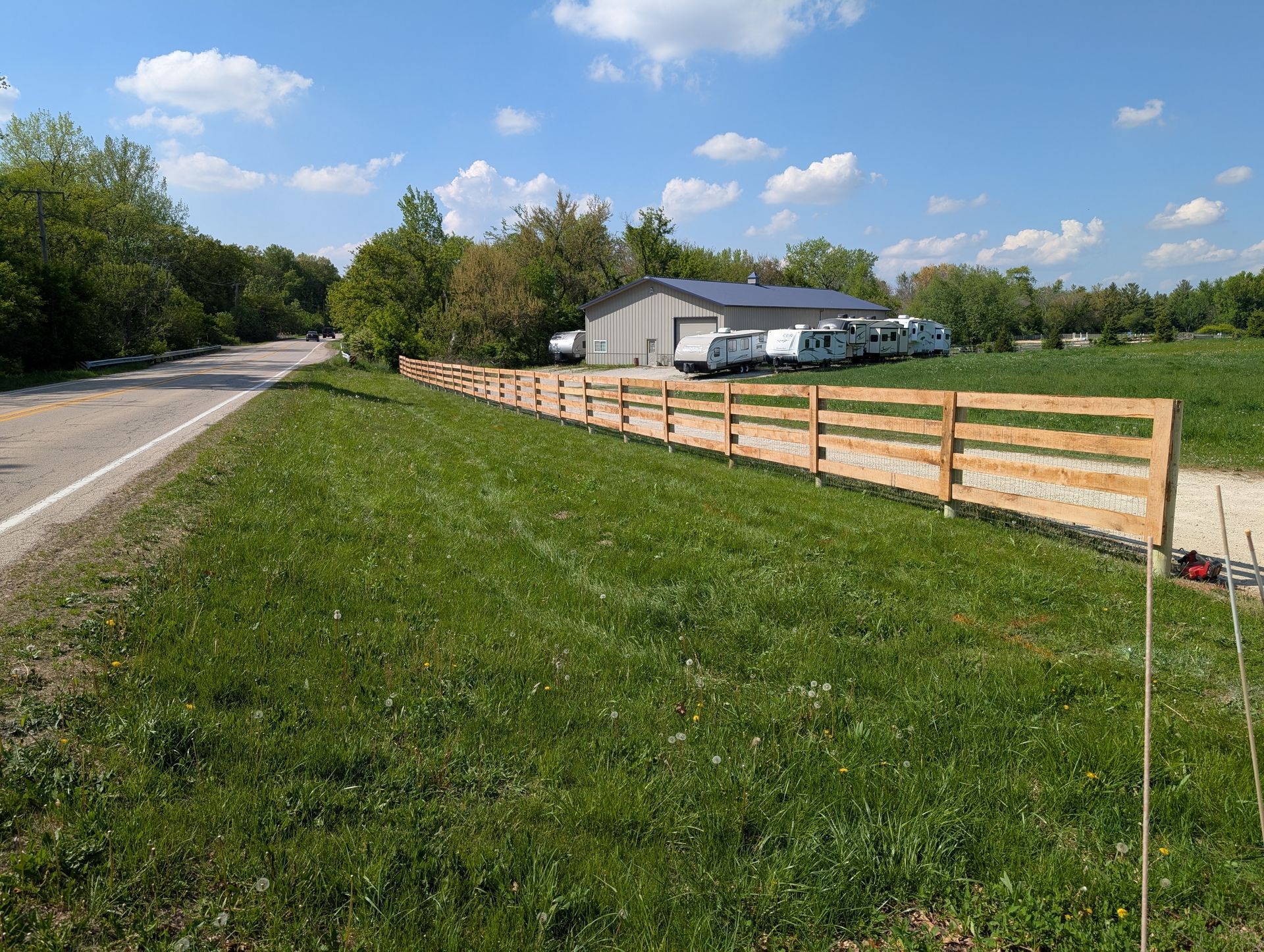 A wooden fence lines a grassy roadside near a metal building storing several campers under a bright blue sky.