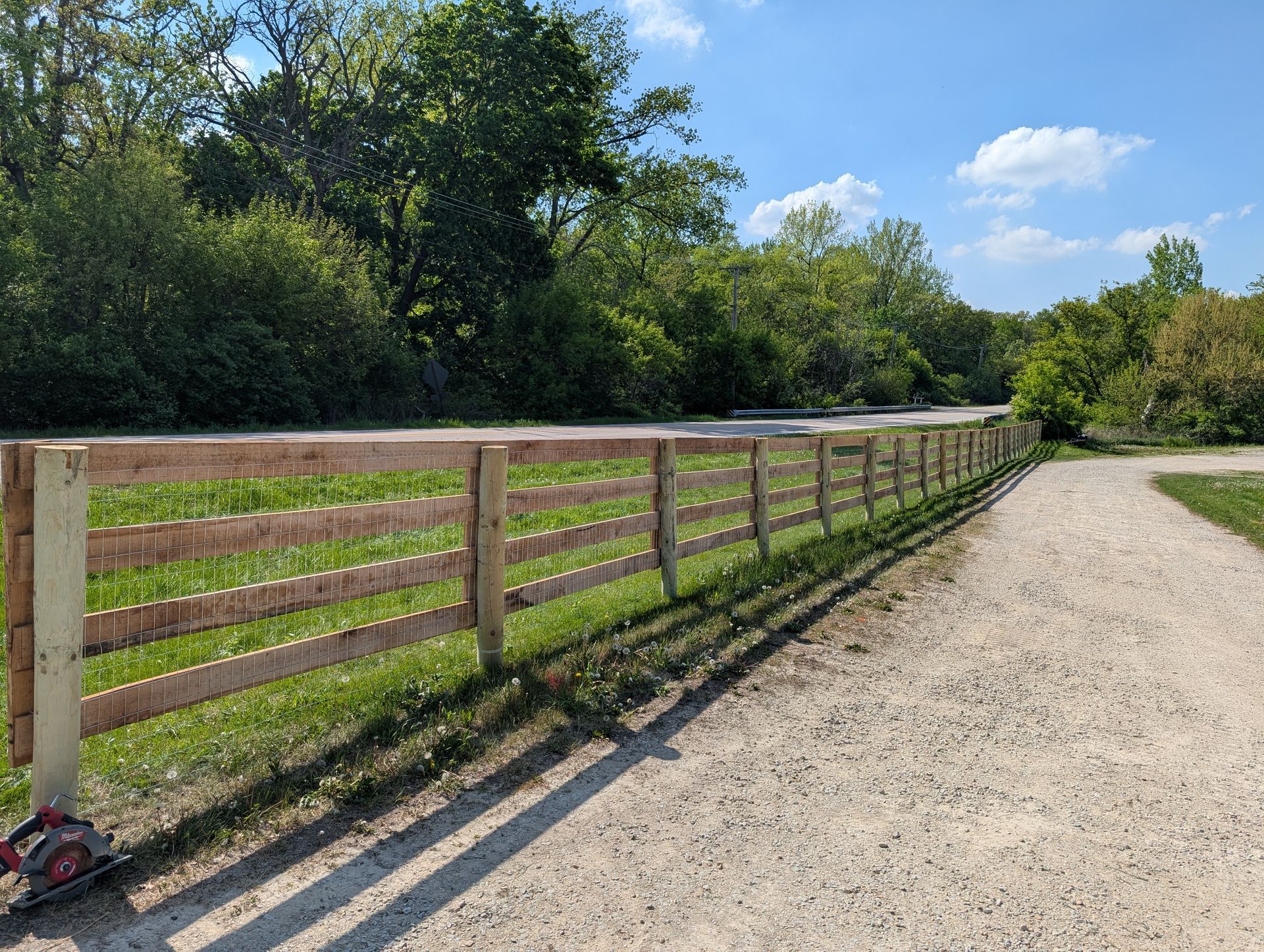 A freshly installed wooden four-rail fence lines a gravel path next to a grassy field and dense woods under a sunny sky.